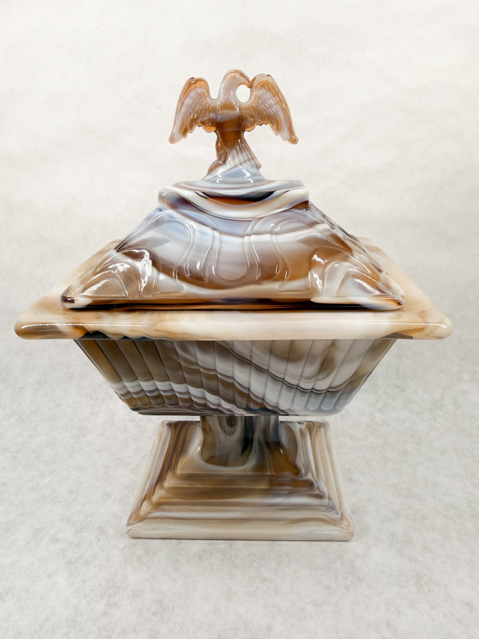 IMPERIAL CARAMEL SLAG COMPOTE, MID-CENTURY: Mid-century caramel slag glass lidded compote produced by Imperial Glass Company featuring a square pedestal form and a spread-wing eagle finial. An interior gold-toned rectangular adhesive label read