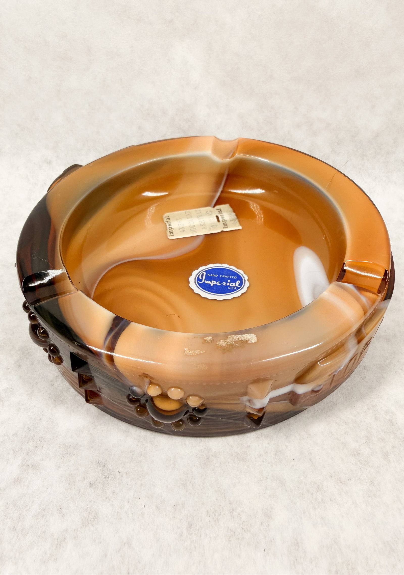 IMPERIAL TOP DAWG SLAG ASHTRAY: An Imperial Glass Company chocolate slag glass ashtray featuring a molded dog collar design with a buckle reading TOP DAWG. The piece retains its original blue scalloped foil label printed HAND