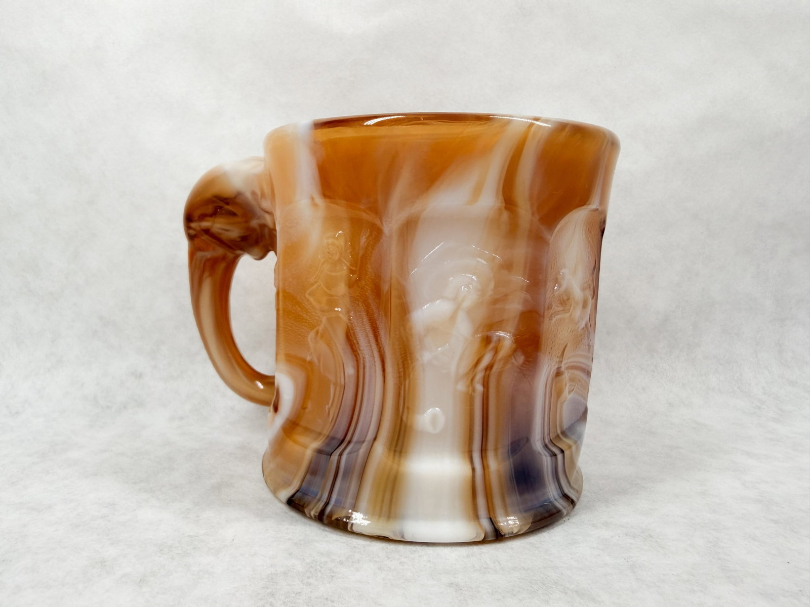 IMPERIAL CARAMEL SLAG GLASS ELEPHANT MUG: A pressed slag glass mug featuring swirled chocolate, caramel, and opaque white coloration with low-relief putti panels and a molded elephant-head handle. The smooth base bears no readable marks or