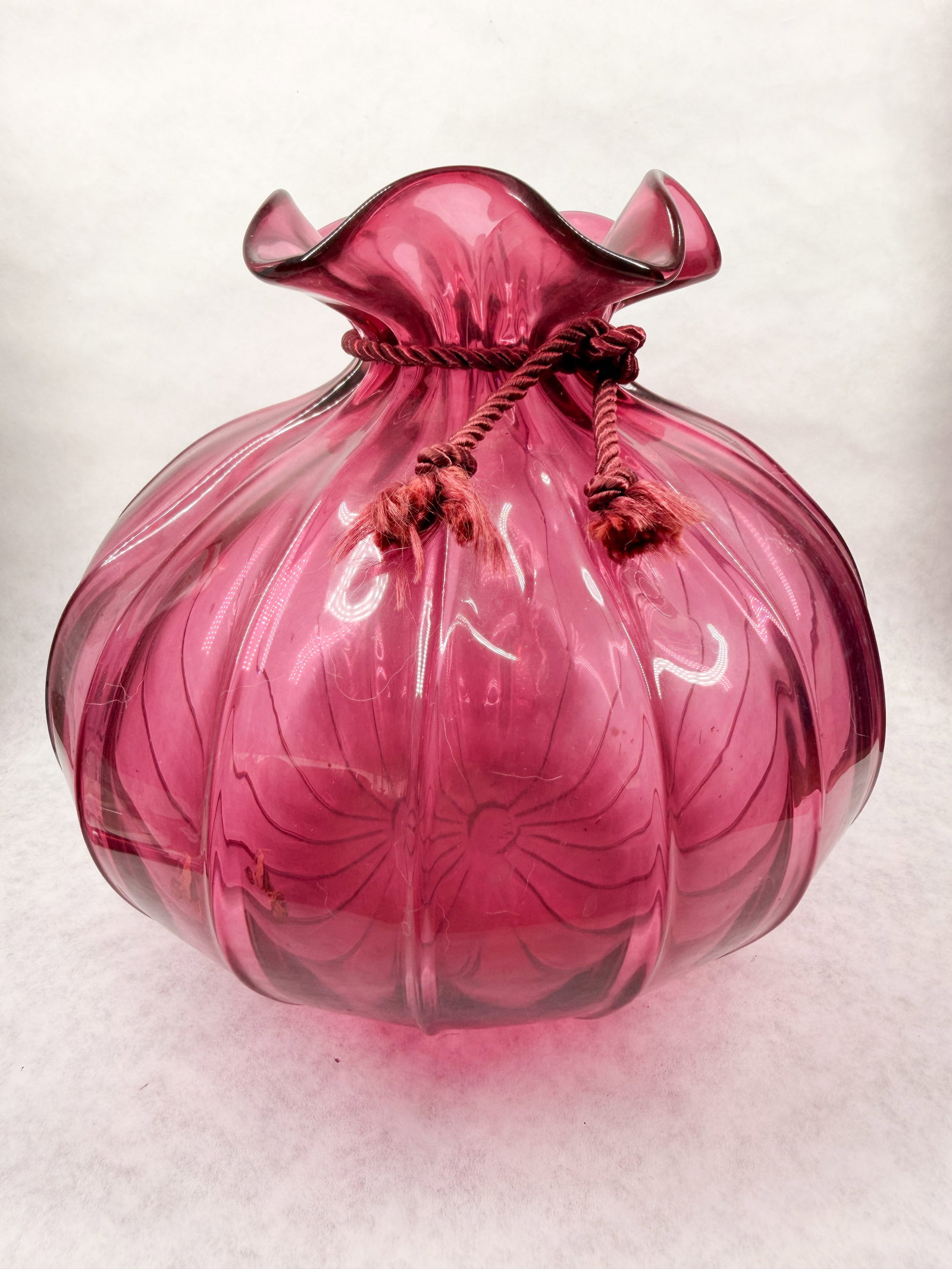 CRANBERRY GLASS RIBBED SACK VASE, 1970S: Hand-blown cranberry glass vase crafted in the form of a cinched drawstring pouch. The vessel features deep vertical ribbing, a ruffled rim, and a rough pontil scar on the underside of the base.