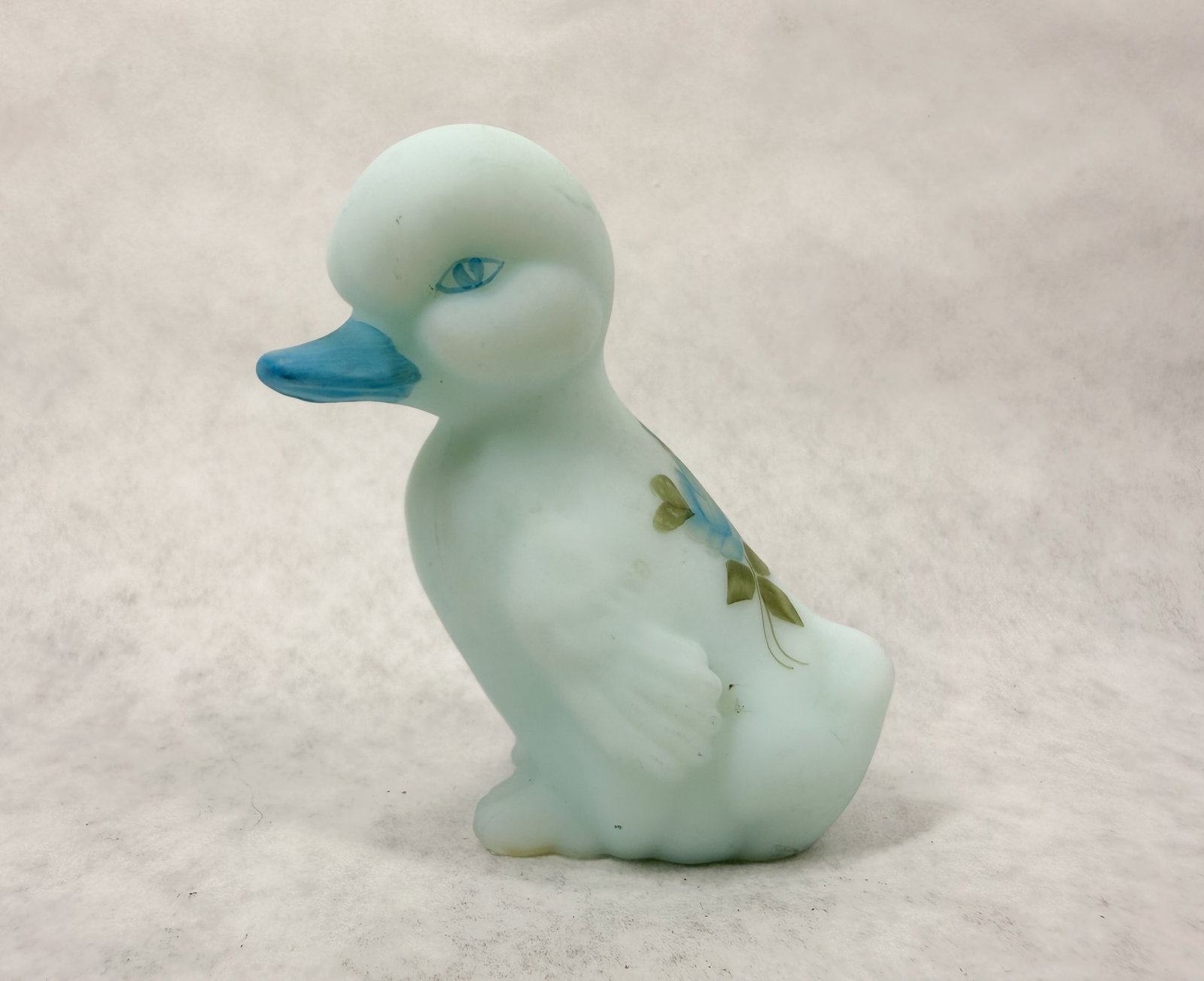 FENTON HP BLUE SATIN GLASS DUCKLING: Seated duckling figurine crafted from light blue satin glass. The underside features a faint, illegible oval mark above a blue script signature reading 'Hand Painted by Lisa G.'.
