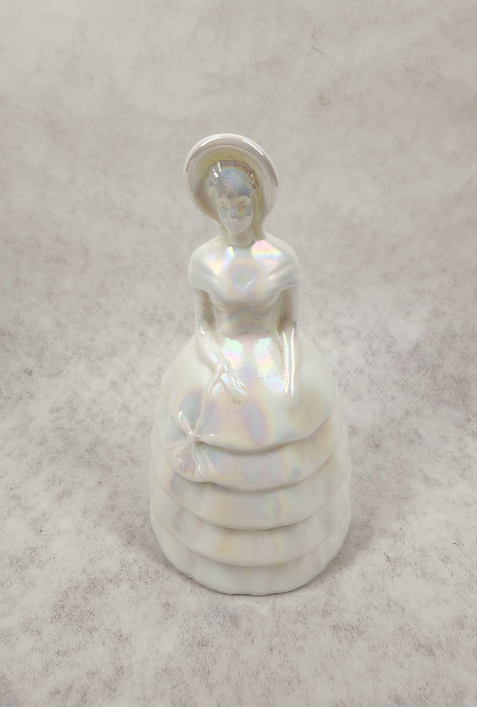 WHITE GLASS SOUTHERN BELLE FIGURINE: Iridescent white glass figurine depicting a Southern Belle in a tiered dress and wide-brimmed bonnet. The piece bears an impressed mark on the lower reverse consisting of a capital letter V within a c