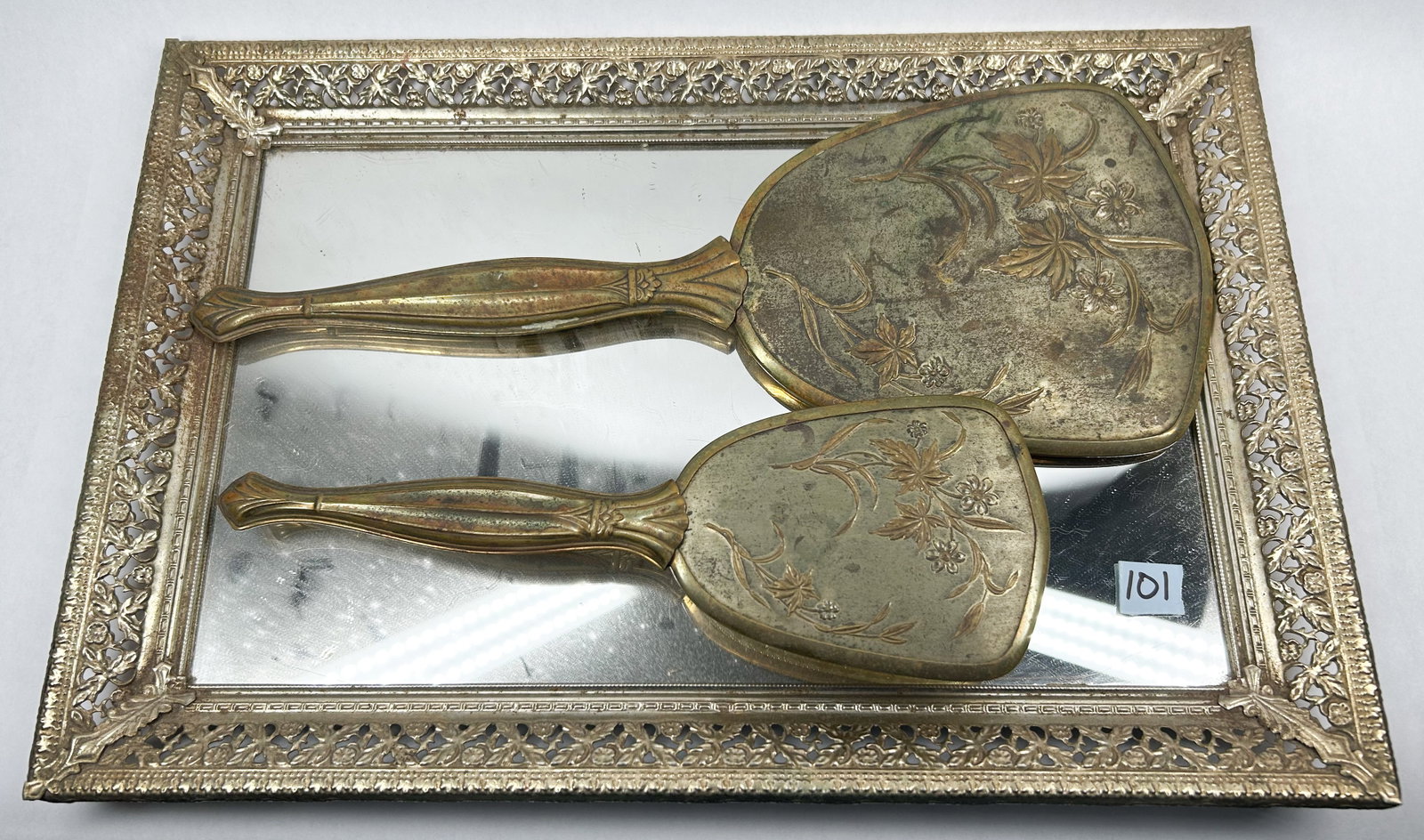 GOLD-TONE THREE-PIECE VANITY SET, CIRCA 1950: A vintage gold-tone metal vanity set comprising a handheld mirror, hairbrush, and rectangular mirrored tray. The pieces feature relief-molded floral motifs and fluted handles; no maker's marks are leg