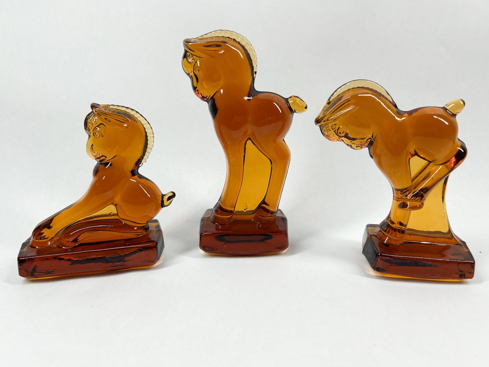 HEISEY BY VIKING AMBER GLASS FOALS: A trio of amber pressed glass Frolicking Foals produced by the Viking Glass Company. Each figure is rendered in a stylized mid-century modern aesthetic and set upon an integral rectangular base. An em