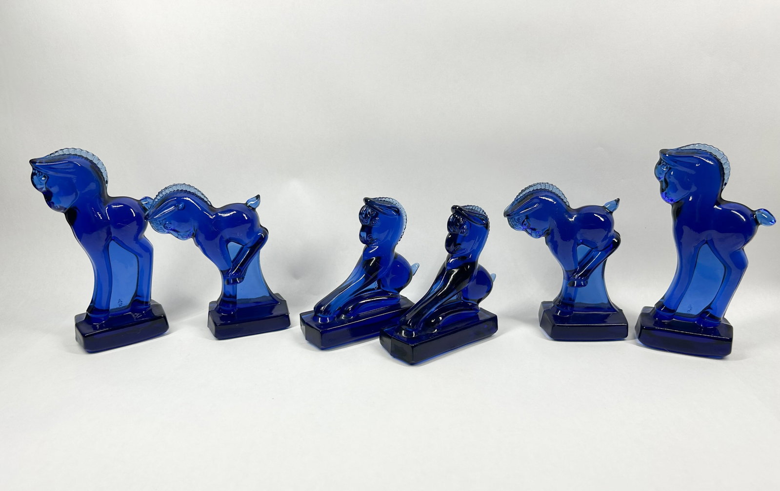 HEISEY BY IMPERIAL ULTRA BLUE COBALT HORSES, C.1960: A set of six translucent cobalt blue glass horse figurines produced by Imperial Glass Company using original Heisey molds. One standing pony retains a blue and silver foil adhesive label on its unders