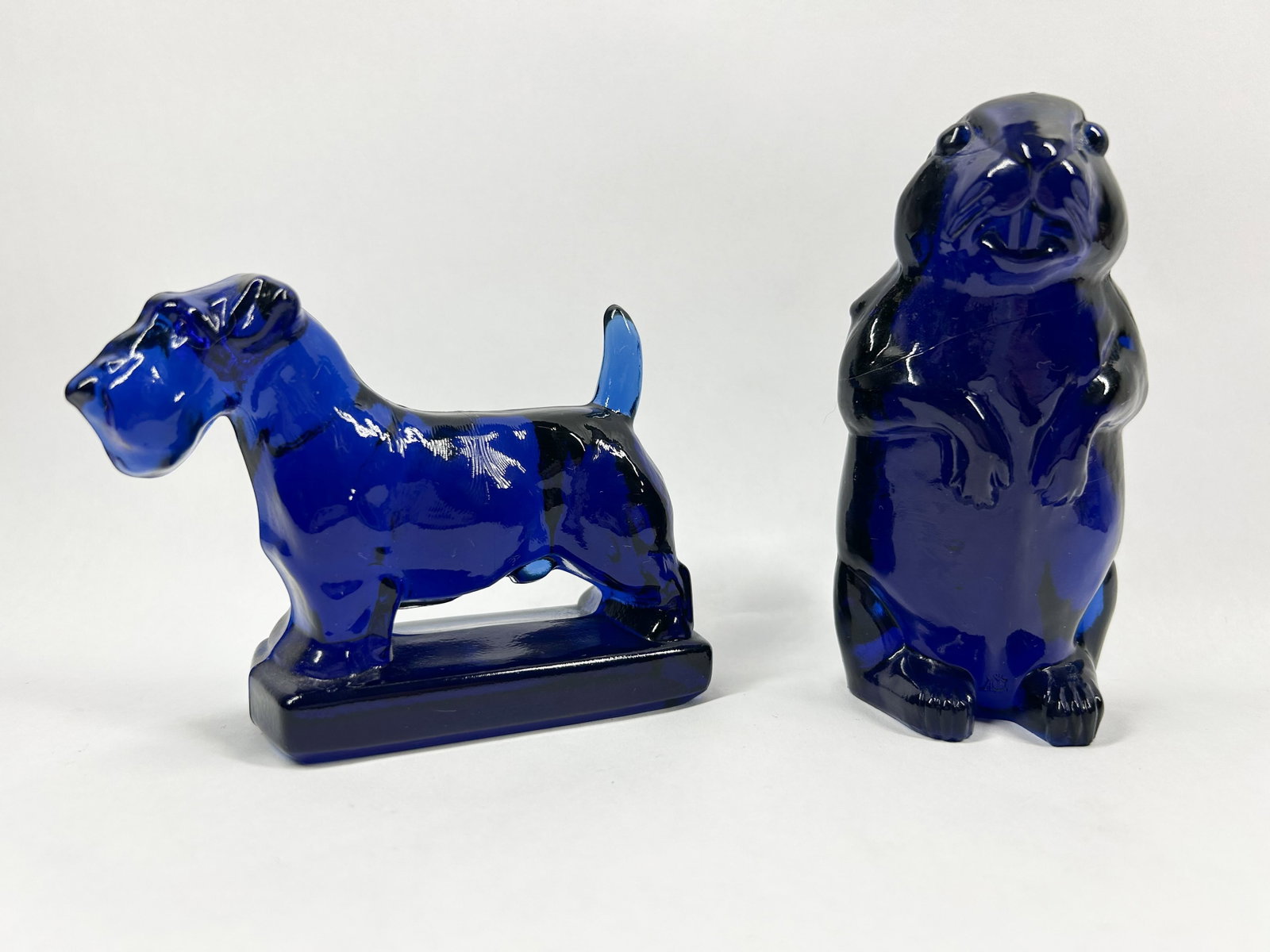 HEISEY BY IMPERIAL ULTRA COBALT BLUE GLASS TERRIER AND WOODCHUCK: A pair of animal figurines comprising a Scottish Terrier on a plinth and a standing groundhog, each fashioned from cobalt blue pressed glass with visible mold seams. The groundhog bears an embossed