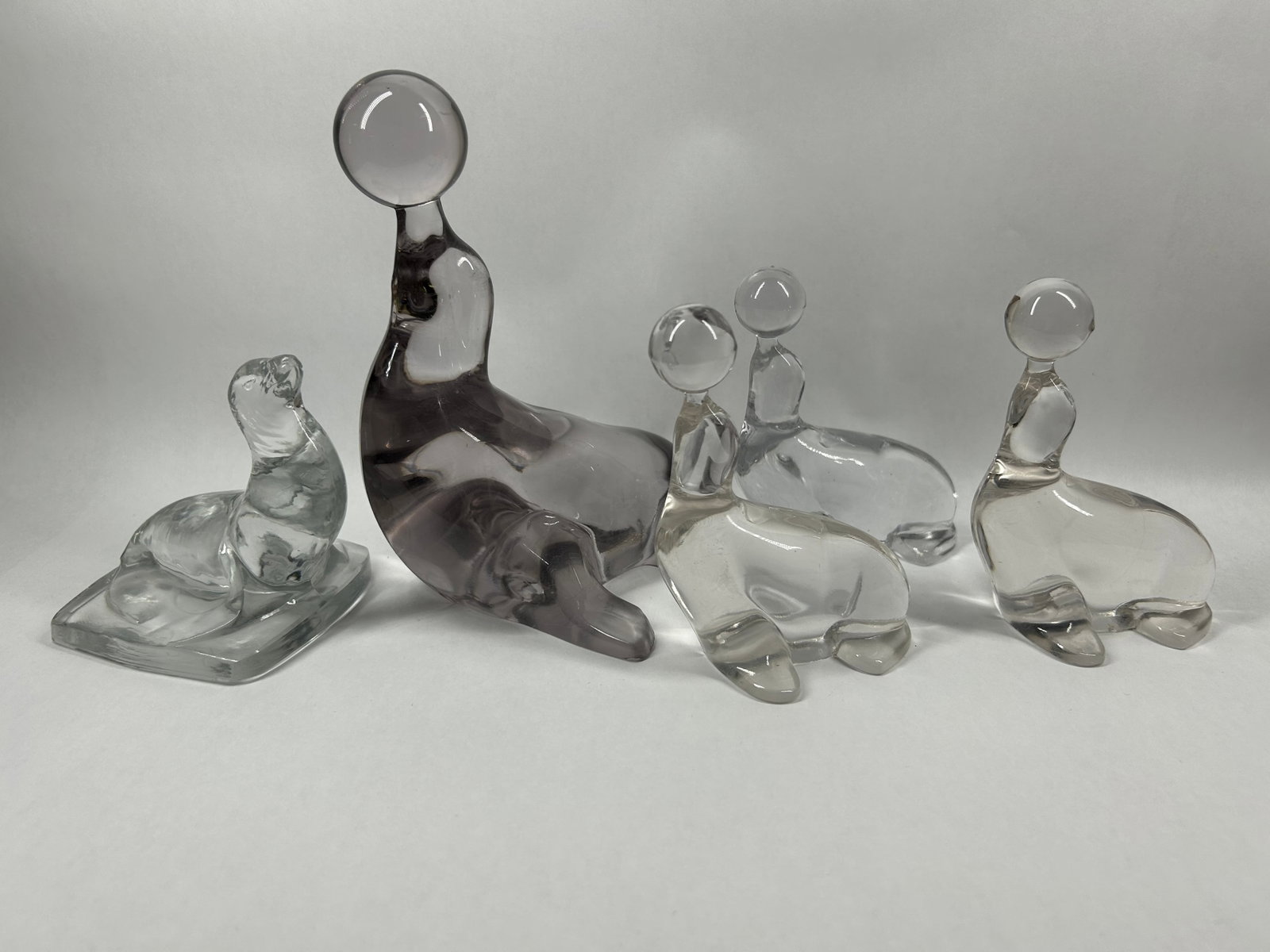 MID-CENTURY GLASS SEAL FIGURINE COLLECTION: A collection of molded glass seal figurines consisting of four stylized forms balancing spheres and one resting figure on a plinth base. One centerpiece is executed in smoke-colored glass, while the