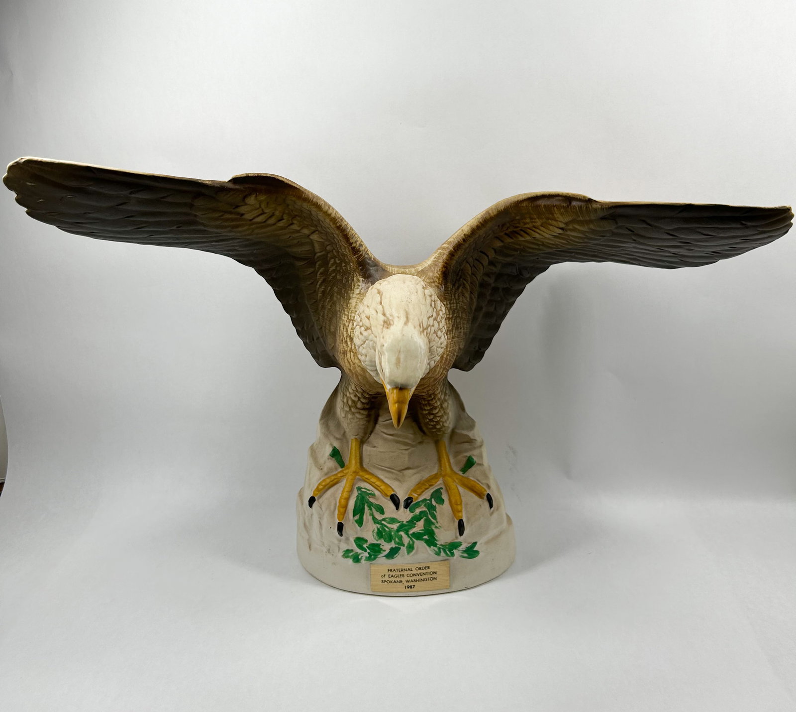 FRATERNAL ORDER OF EAGLES CERAMIC DECANTER, 1987: Hand-painted ceramic commemorative decanter modeled as a bald eagle with outspread wings perched on a rock-like base with green foliage. A gold-toned rectangular adhesive label on the base is printed 