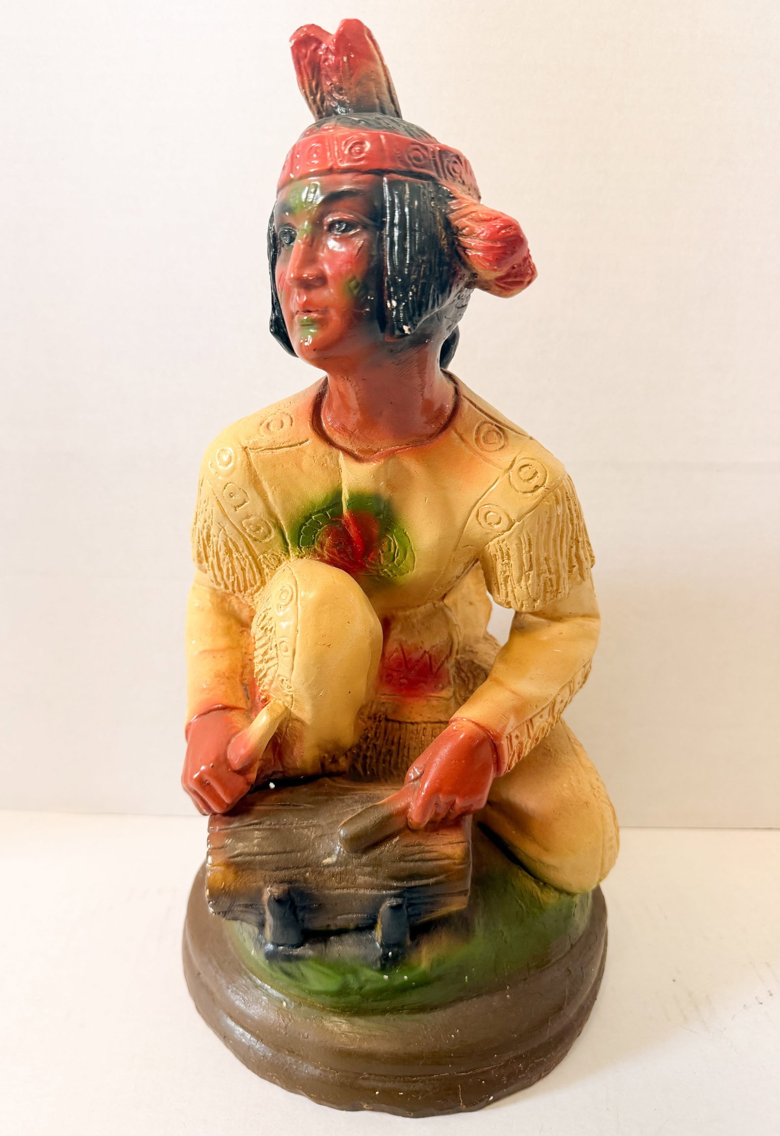 CHALKWARE NATIVE AMERICAN DRUMMER, MID-20TH CENTURY: A mid-20th-century hand-painted chalkware figurine depicting a kneeling Native American figure playing a drum. The figure is decorated with a fringed tan tunic and a red feathered headband on a