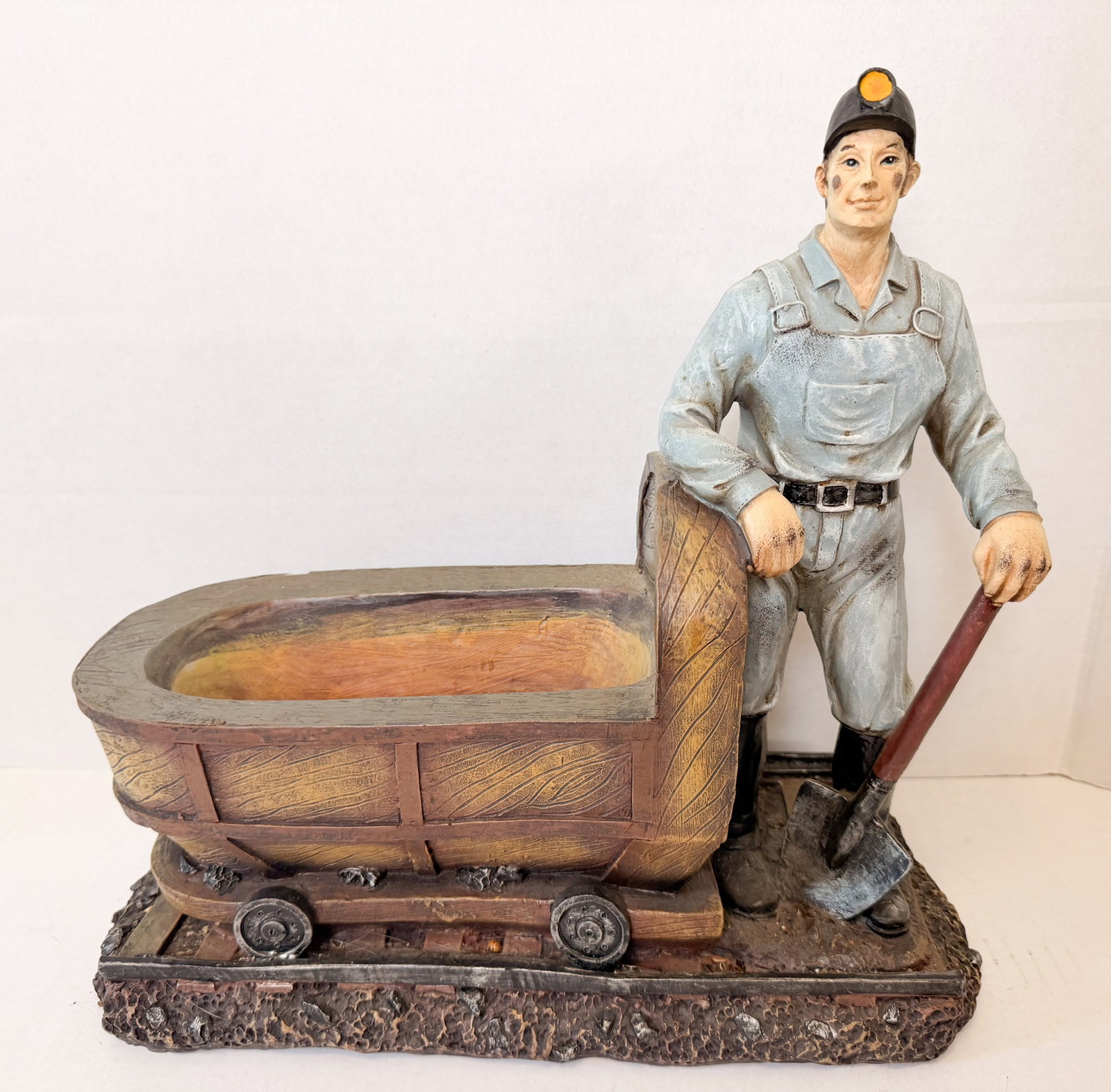 RESIN COAL MINER PLANTER FIGURINE: Cast resin figurine depicting a standing coal miner with a shovel beside a hollow mine cart on a railroad track base. The piece features realistic painted textures and simulated weathering with no