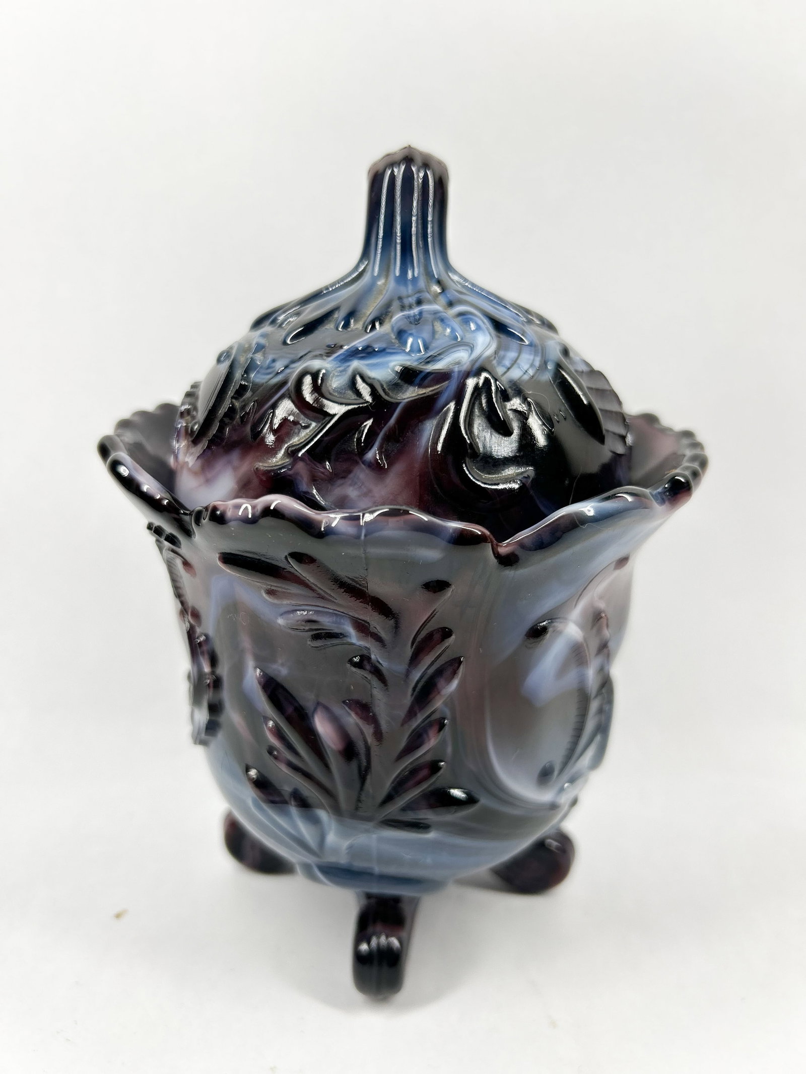 IMPERIAL PURPLE SLAG GLASS LIDDED BOWL: A three-footed lidded vessel crafted in purple and white swirled pressed slag glass. The exterior features relief-molded acanthus leaves and scrolls with a scalloped rim and a ribbed finial. No maker 