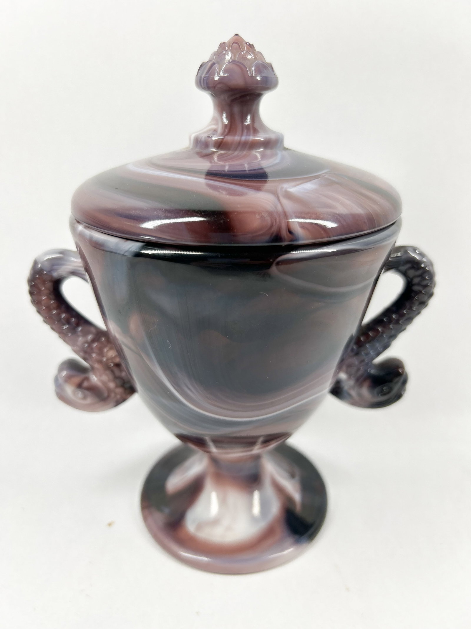 PURPLE SLAG GLASS SUGAR BOWL: A covered sugar bowl in purple and white swirled pressed slag glass. The urn-shaped vessel features dual figural handles and a matching lid with a textured finial.