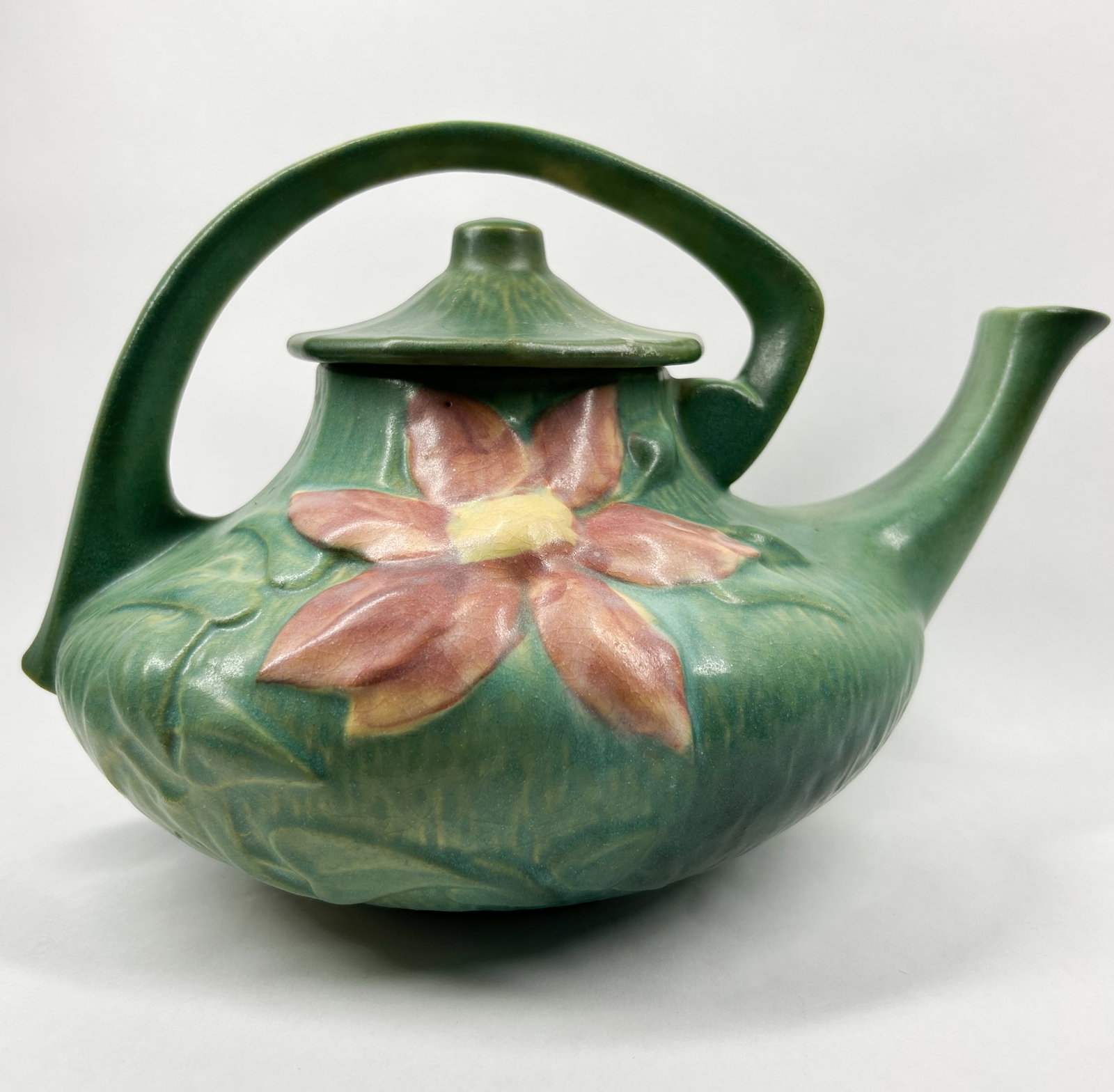 ROSEVILLE GREEN FLORAL EARTHENWARE TEAPOT: An American art pottery earthenware teapot featuring embossed floral motifs on a textured green ground. The piece retains its original conical lid and displays an integral high-arching handle.