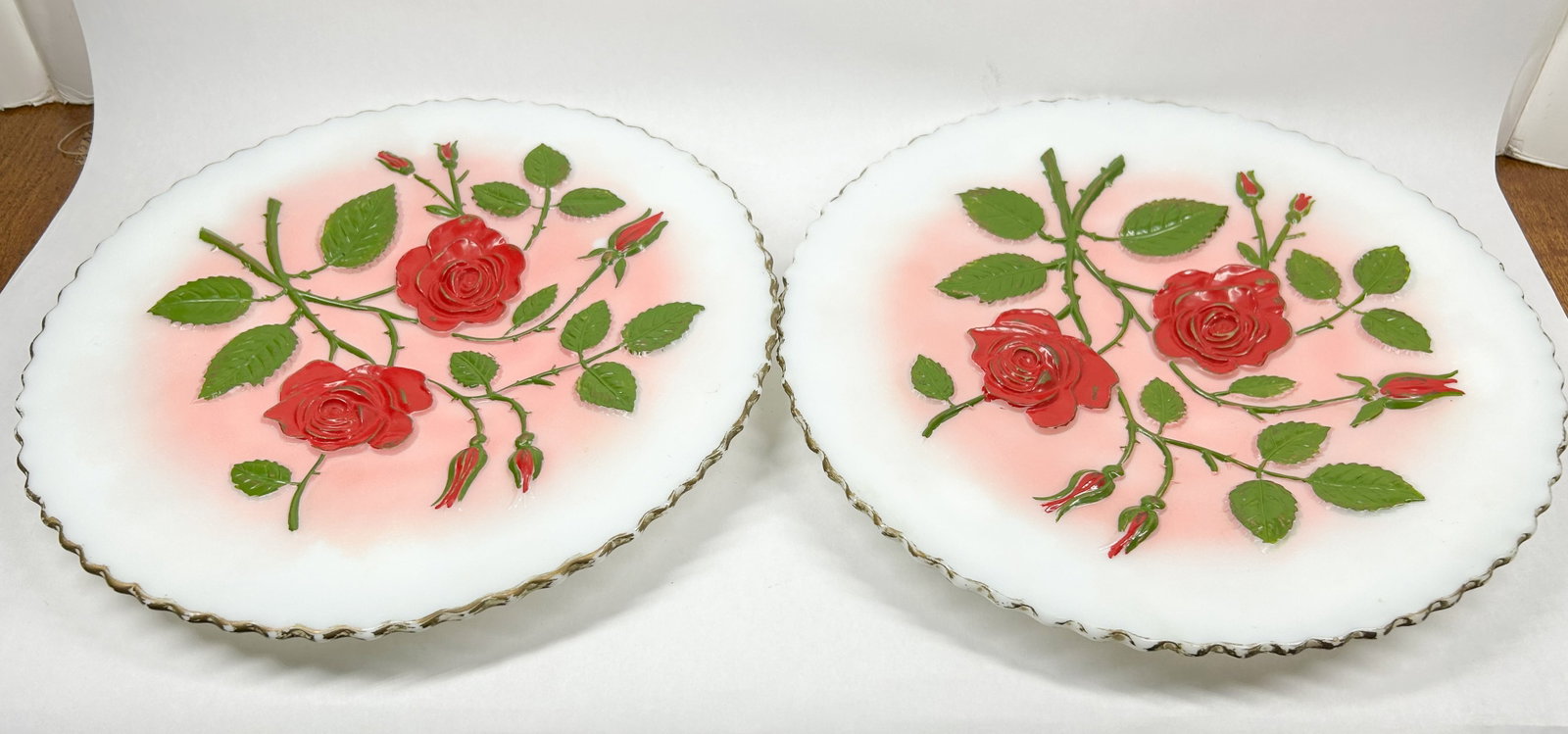 CAMBRIDGE MILK GLASS CHARGERS: A pair of white milk glass chargers featuring high-relief rose and foliage decoration finished with polychrome hand-painted enamel and gilded scalloped rims. A molded rose device is centered on the un