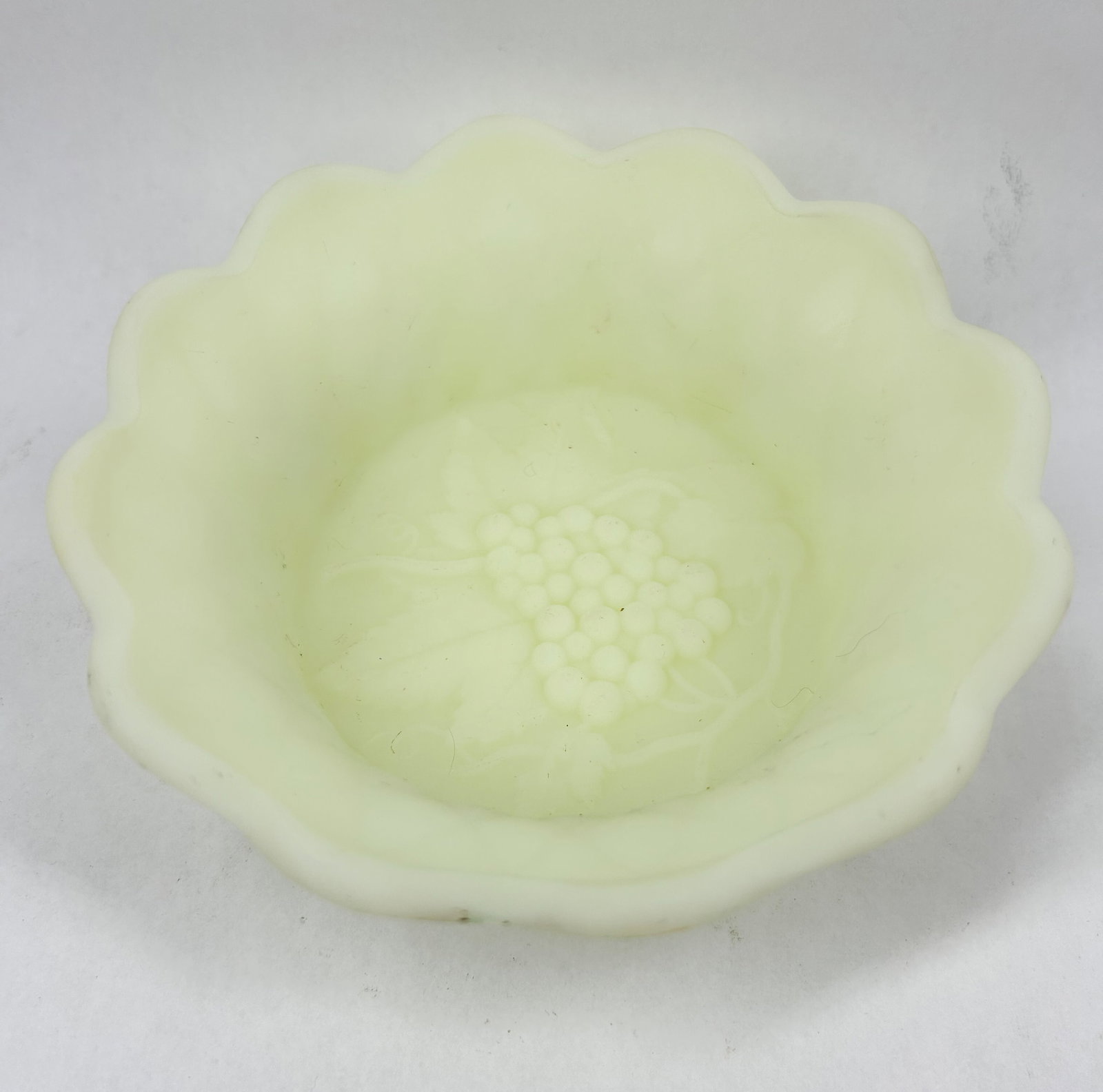 CUSTARD GLASS GRAPE BOWL, CIRCA 1910: A pale yellow-green custard glass dessert bowl featuring a satin finish and an embossed grape motif, with an impressed capital letter N in a circle centered within a sunburst pattern on the underside 