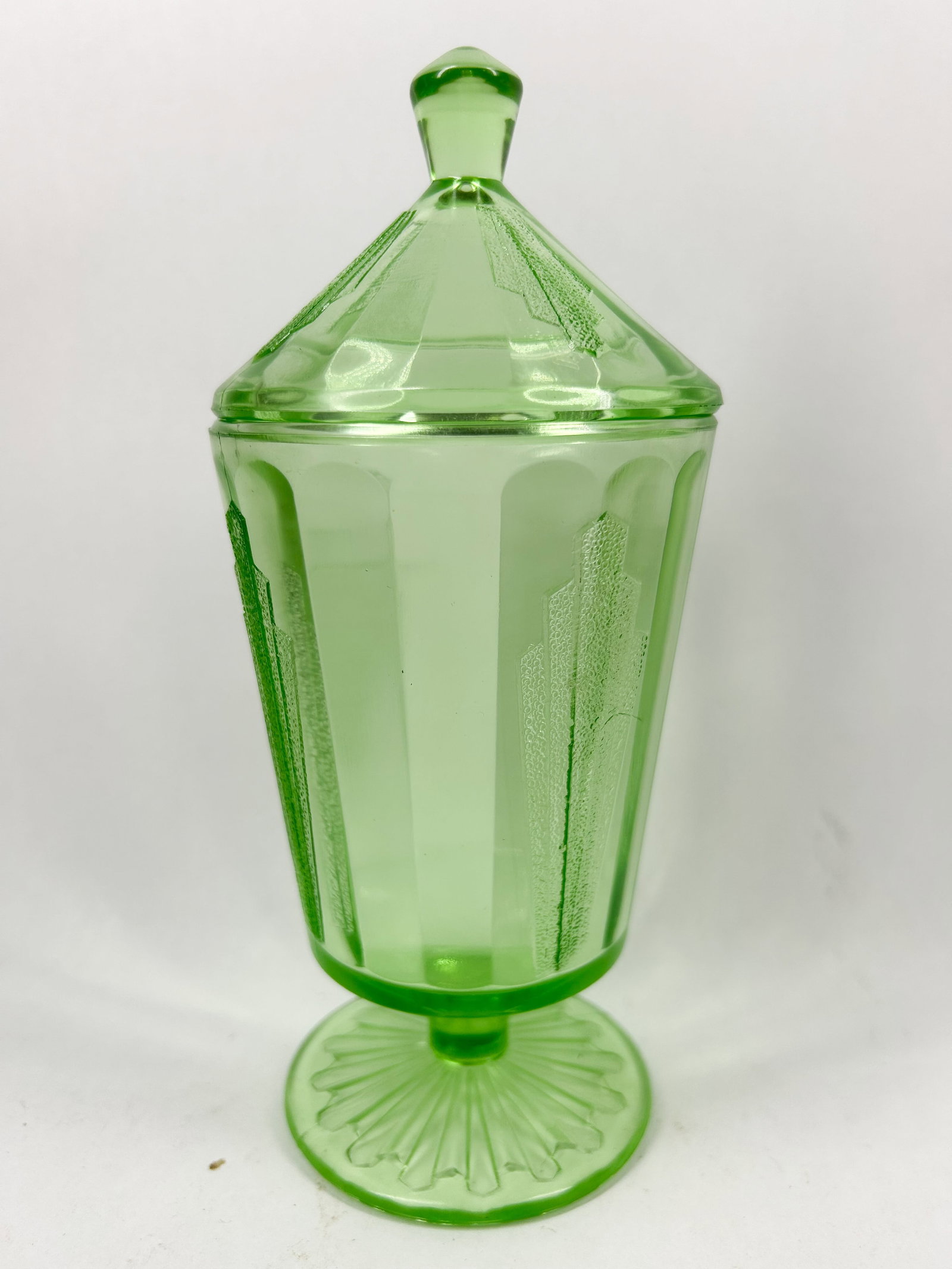 GREEN URANIUM GLASS LIDDED JAR, C. 1930: A pressed green uranium glass pedestal jar and matching lid featuring stylized Art Deco architectural motifs. The tapered body and domed lid display a stippled texture and sit upon a circular foot wit