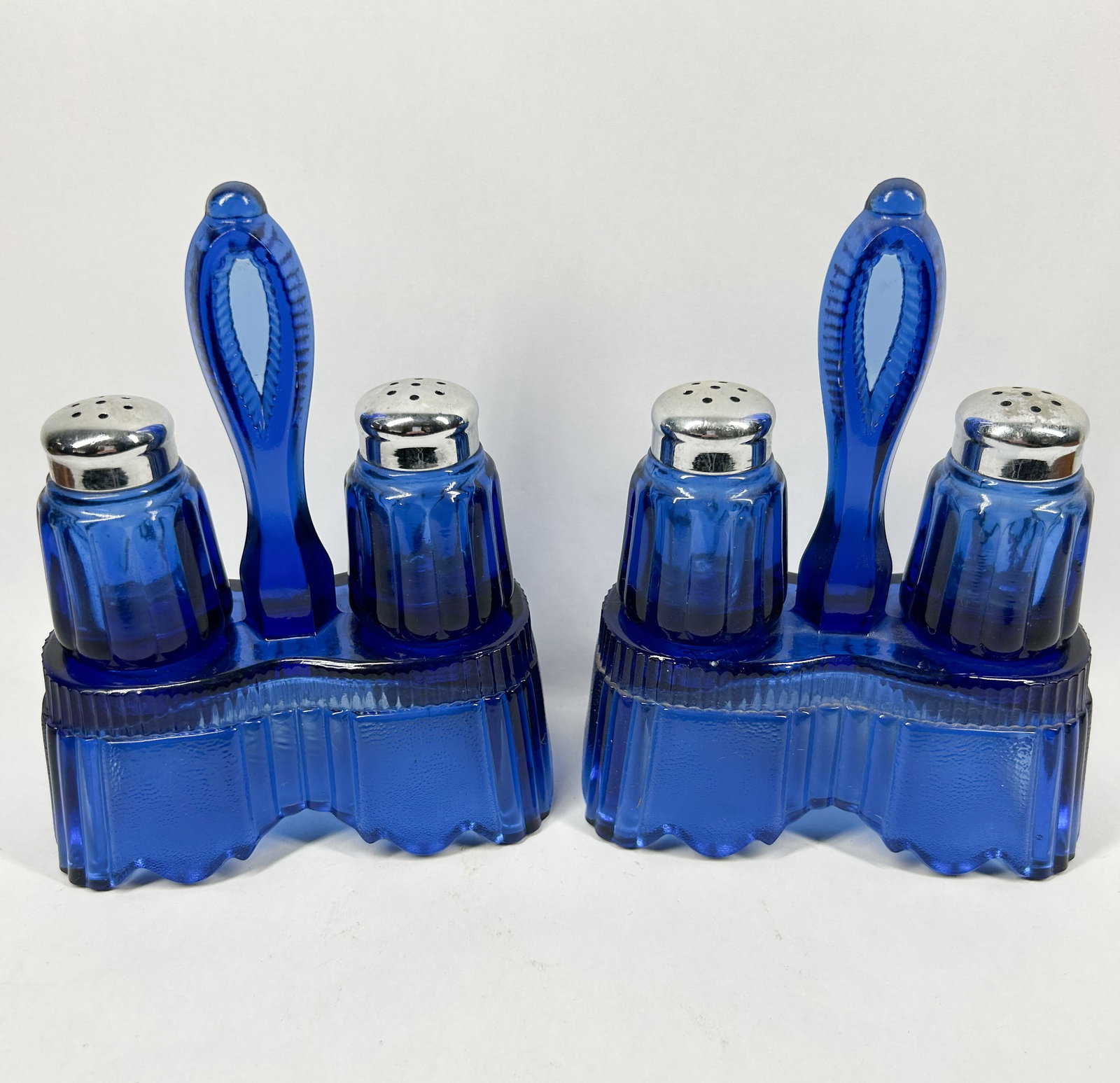 PAIR OF COBALT GLASS CASTER SETS, CIRCA 1930: A pair of cobalt blue glass salt and pepper caster sets. The molded glass features a vertical ribbed pattern and is fitted with silver-toned perforated lids.