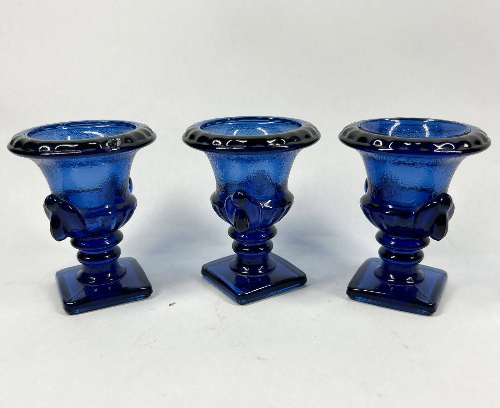 COBALT BLUE GLASS MINIATURE URNS: A trio of cobalt blue pressed glass miniature urns. The vessels feature gadrooned rims and twin scroll handles above stepped square pedestal bases.