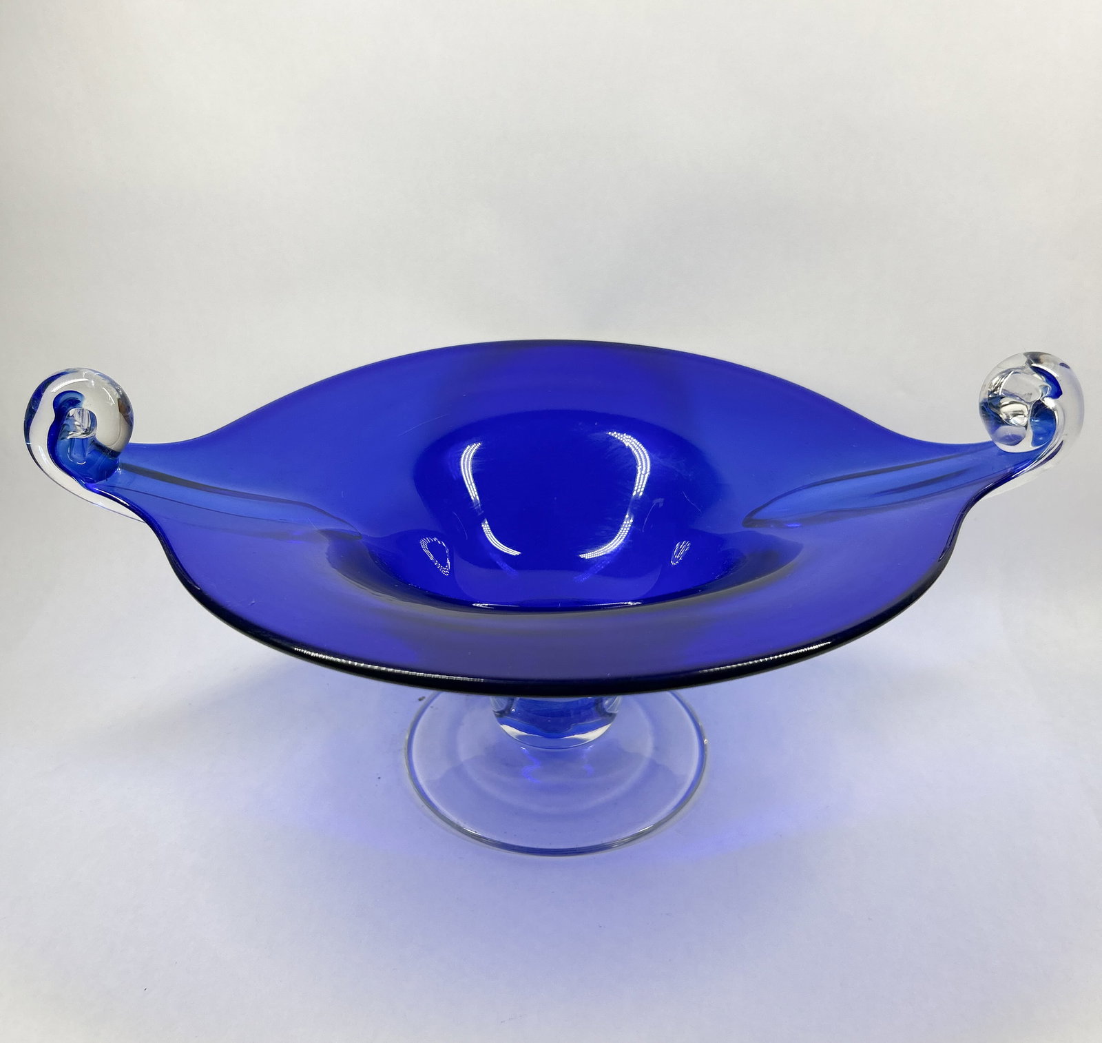 COBALT ART GLASS PEDESTAL BOWL: Hand-blown cobalt blue glass bowl featuring clear glass scrolled handles and a pedestal base with a compressed knop. The construction includes applied hot-worked glass elements consistent with