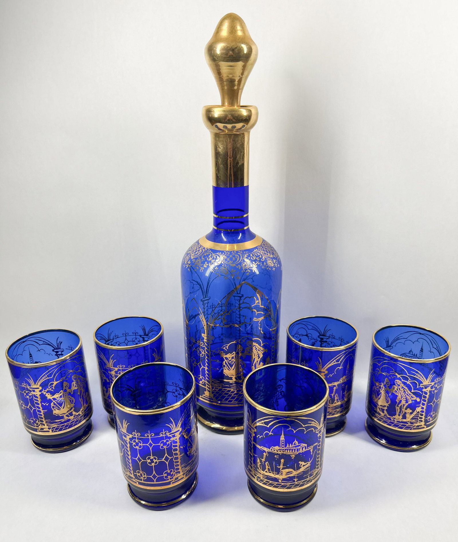 SEVEN-PIECE COBALT VENETIAN GLASS SET, MID-20TH CENTURY: A seven-piece set of translucent cobalt blue glassware comprising a decanter with a flame-shaped gilded stopper and six matching tumblers. Each piece is decorated with hand-painted gold enamel