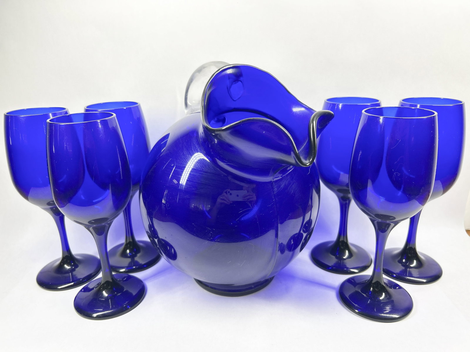 COBALT BLUE GLASS BEVERAGE SET: Seven-piece beverage set consisting of a bulbous cobalt blue glass pitcher with a ruffled rim and applied clear handle, together with six matching wine glasses. The glasses feature rounded bowls, inte
