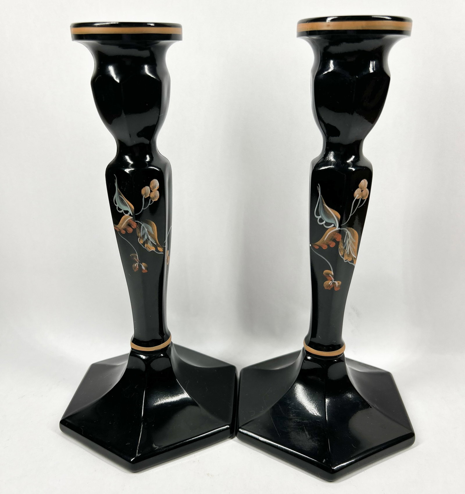 FENTON PAINTED BLACK GLASS CANDLESTICKS: A pair of opaque black glass candlesticks featuring hexagonal bases and faceted baluster stems. The pieces are decorated with hand-painted floral motifs and gold-toned accents.