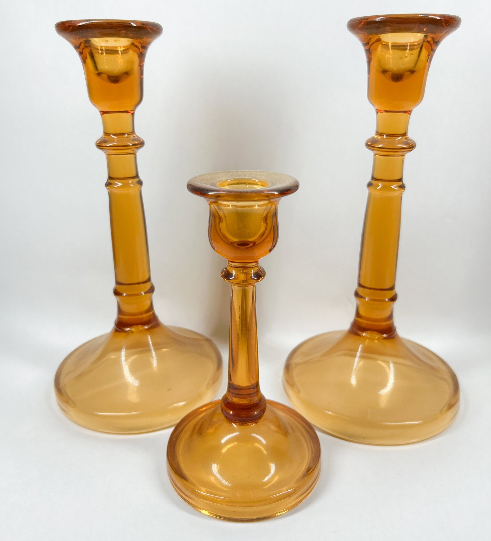 AMBER GLASS CANDLESTICK TRIO, MID-20TH CENTURY: A set of three amber glass candlesticks featuring tapered stems with three horizontal knops and wide circular domed bases. The collection consists of a matching pair and one shorter coordinating