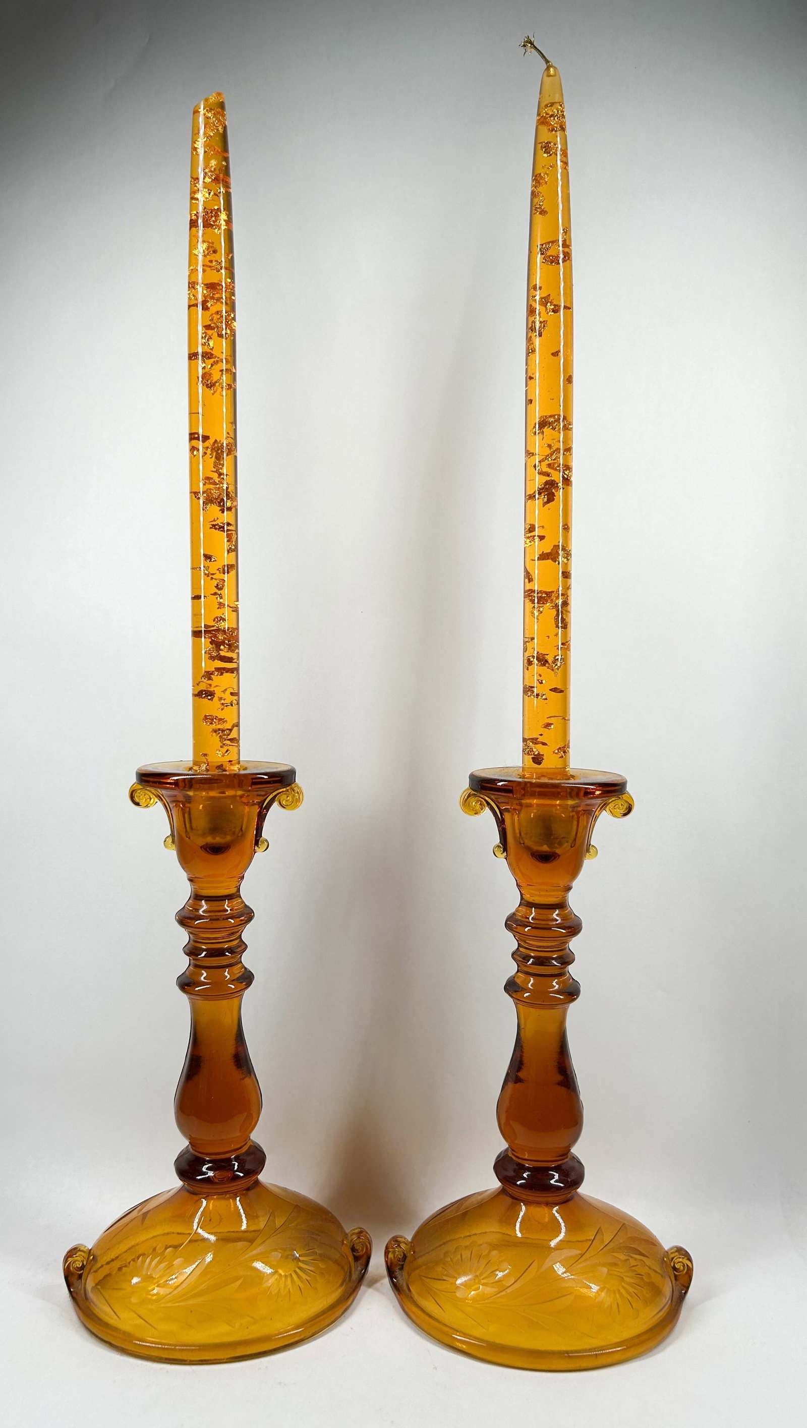 AMBER GLASS CANDLESTICKS AND OIL CANDLES, MID-CENTURY: A pair of amber glass candlesticks featuring floral-etched domed bases with scrolled tabs, paired with matching glass oil candles containing suspended gold-toned flakes. No signatures, labels, or