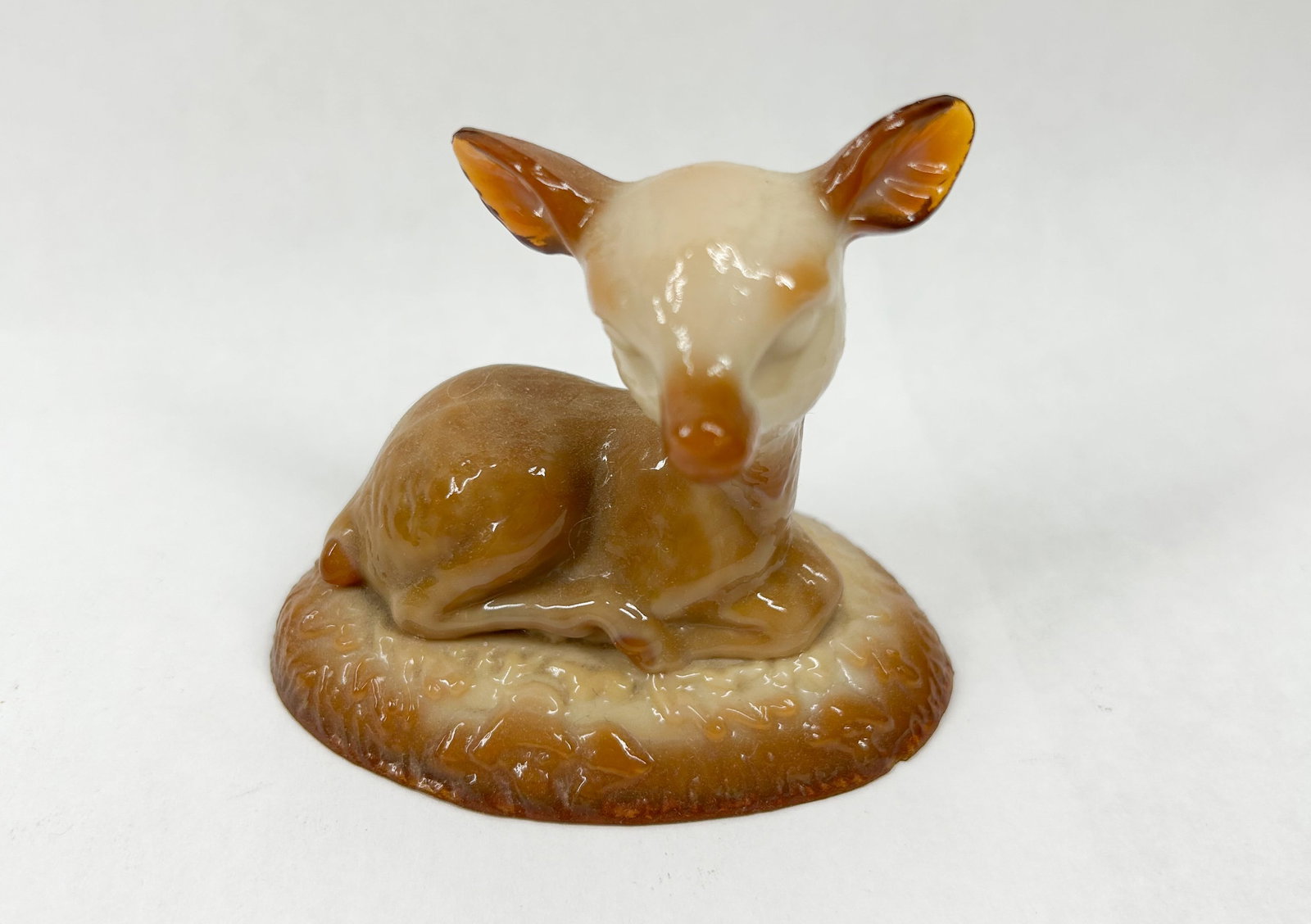 BOYD BINGO DEER CARAMEL SLAG: A small figurine depicting a reclining fawn, crafted from variegated caramel slag glass. The figure is posed on an integral oval base featuring a molded, textured surface representing grass.
