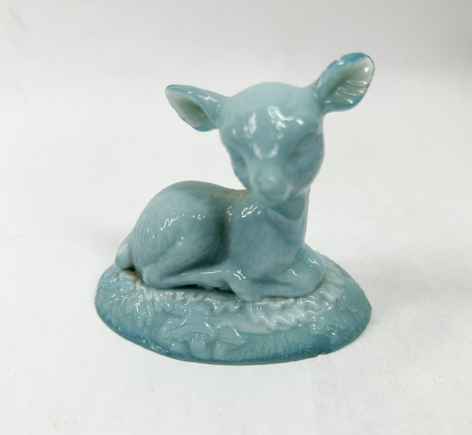 BOYD LIGHT BLUE BINGO DEER: A decorative figurine depicting a reclining fawn in swirled blue and white slag glass. The figure is modeled in a resting pose atop an oval, textured base simulating grass.