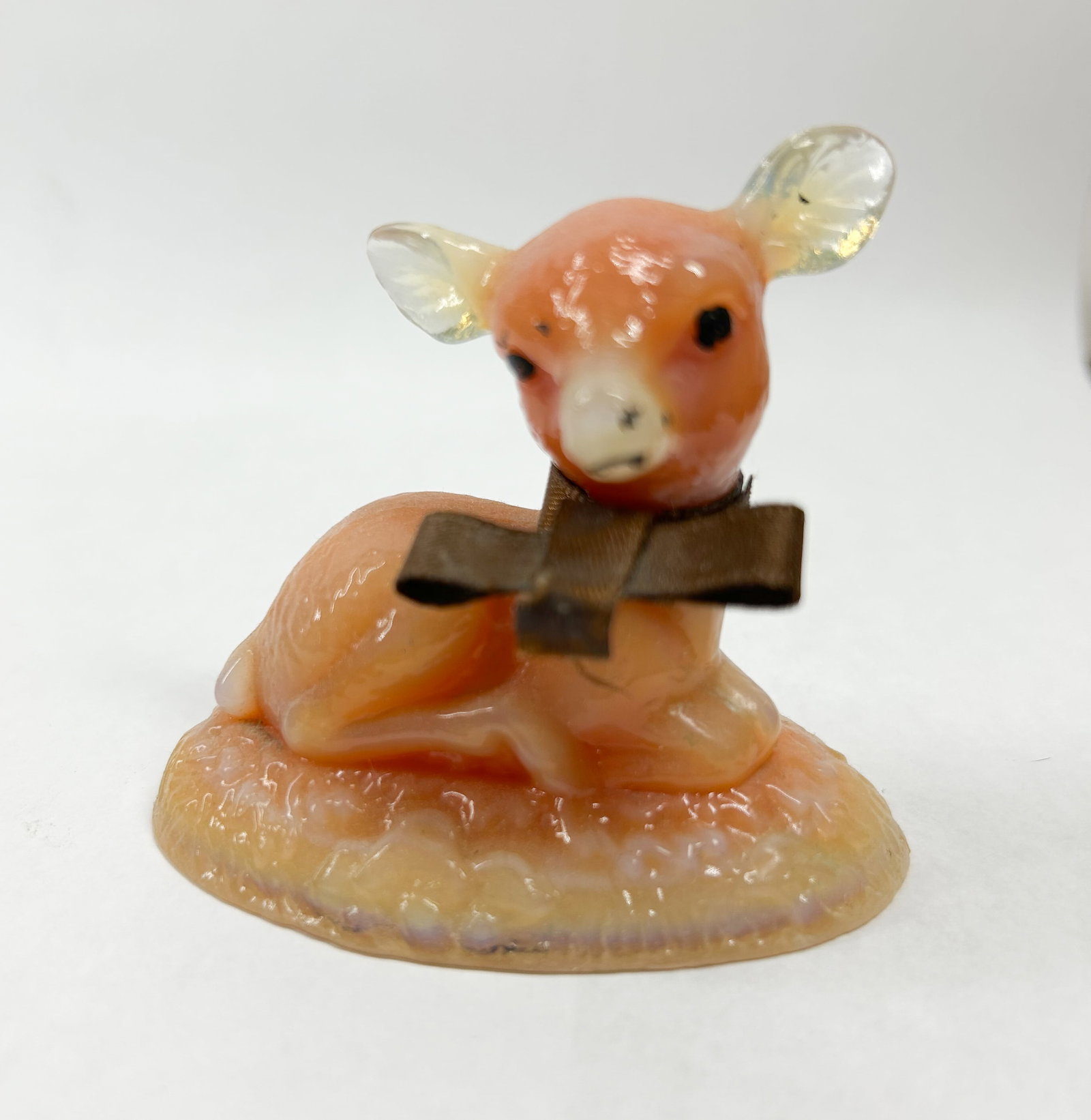BOYD PEACH BINGO DEER: Molded slag glass figurine of a reclining fawn featuring peach and cream marbling. The ears and textured base exhibit opalescent properties, and the neck is adorned with a brown textile ribbon.