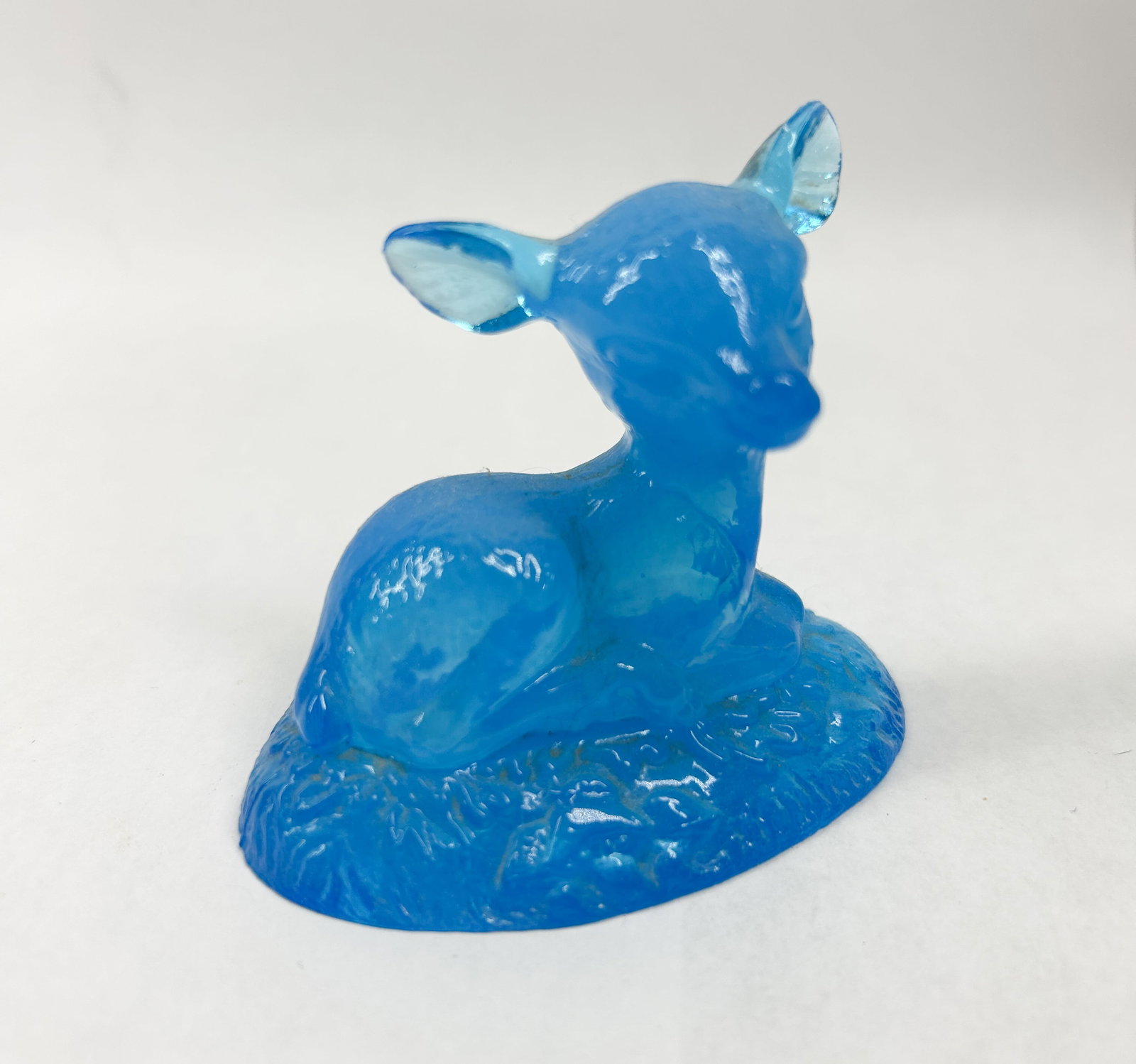 BOYD BLUE BINGO DEER: A translucent blue glass figurine depicting a reclining fawn on a textured base. The piece is crafted from molded glass with vibrant color and subtle internal variations.