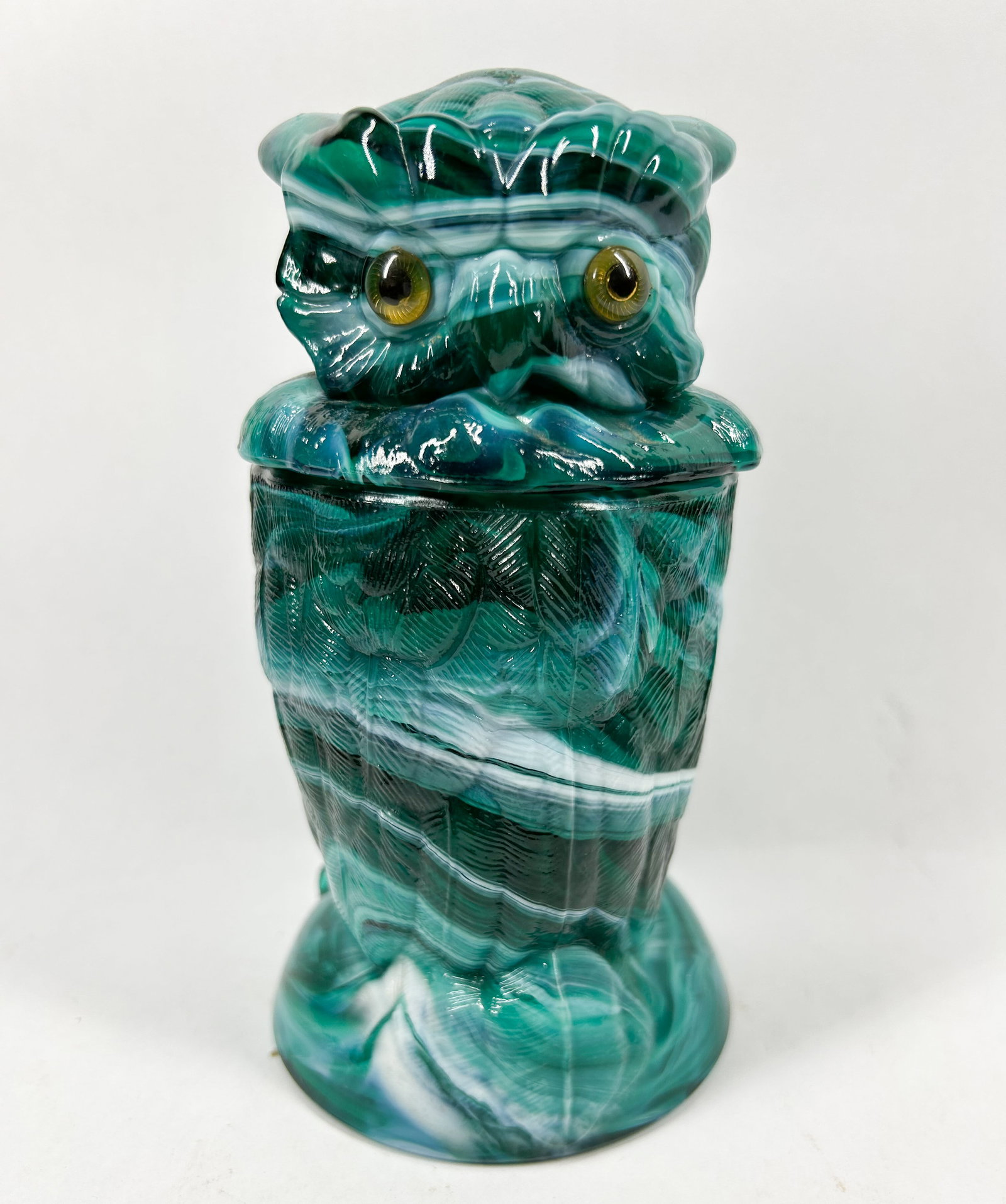 IMPERIAL GREEN SLAG GLASS OWL JAR: A lidded jar modeled in the form of a perched owl, composed of forest green and white swirled slag glass. The two-piece vessel features relief-molded plumage, a flared circular base, and two inset amb
