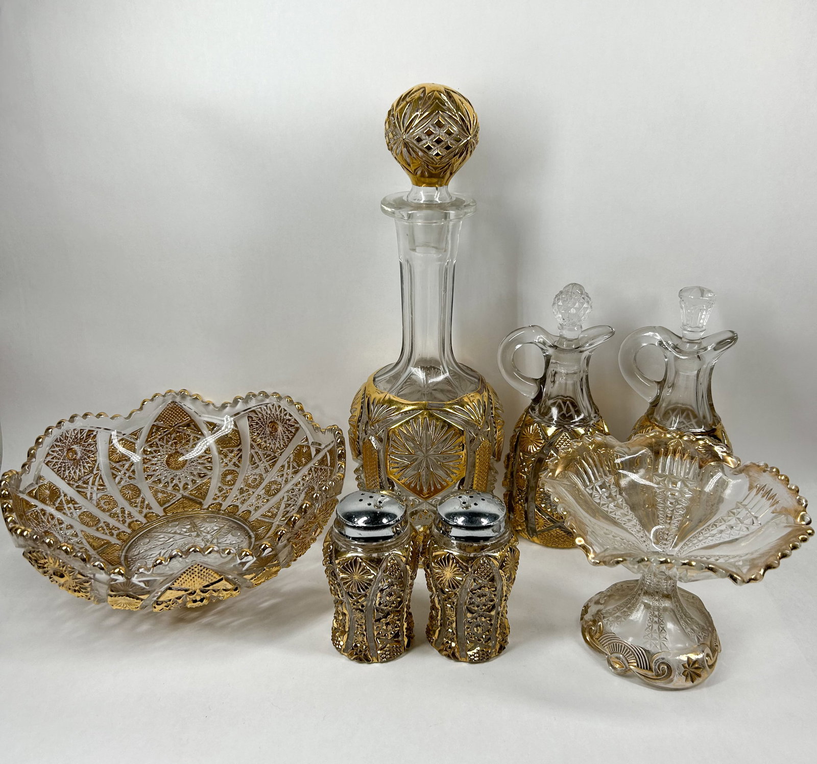 SEVEN-PIECE GILT PRESSED GLASS SERVICE, MID-20TH CENTURY: A matching seven-piece table service crafted from clear pressed glass featuring faceted starburst patterns and heavy gold-gilt highlights. The collection comprises a centerpiece bowl, a decanter with 