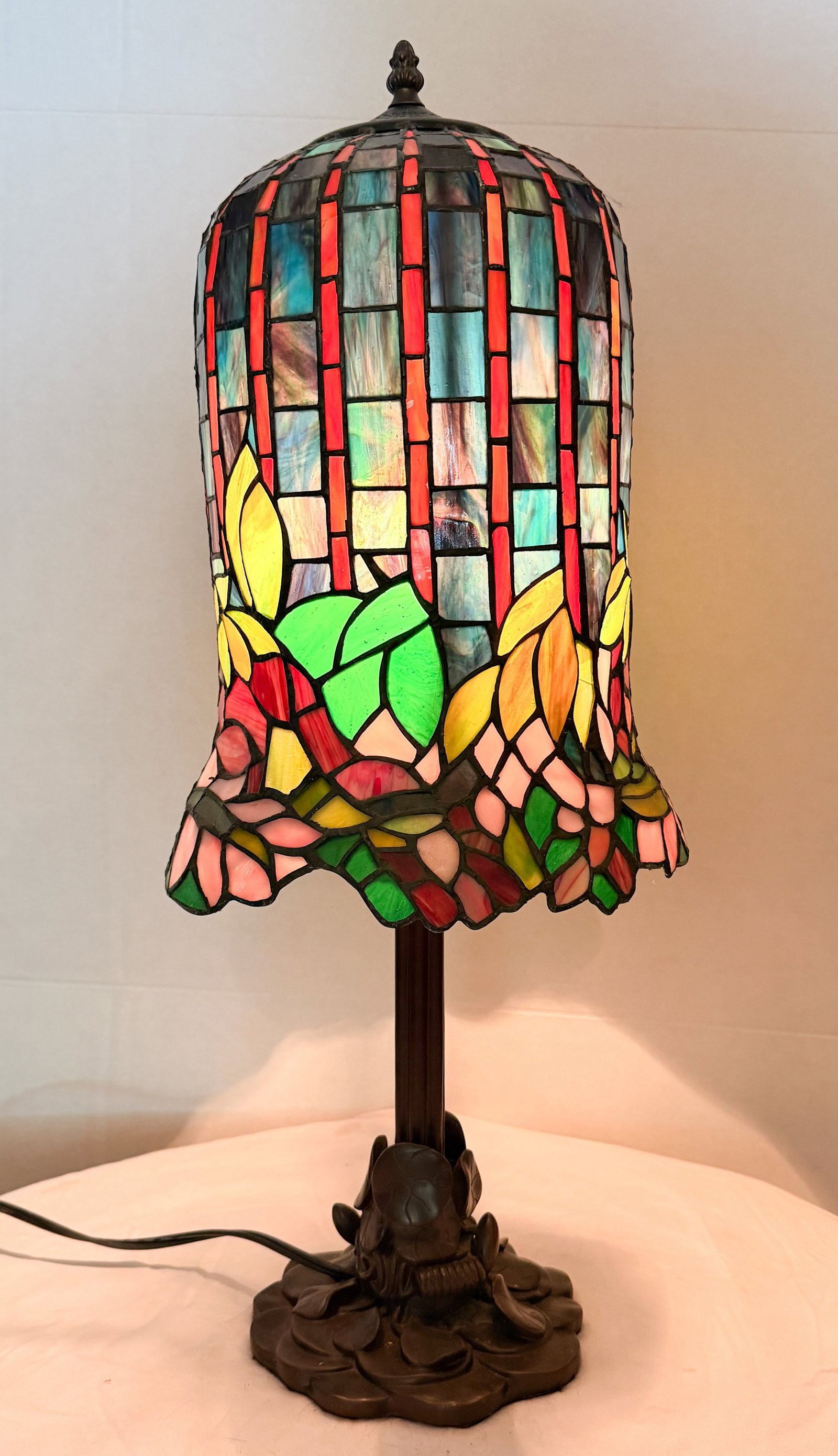 TIFFNY STYLE POND LILY TABLE LAMP: A leaded glass table lamp featuring a bell-shaped shade with a geometric upper register and floral frieze. The shade is supported by a dark brown patinated cast metal base with an overlapping lily pad