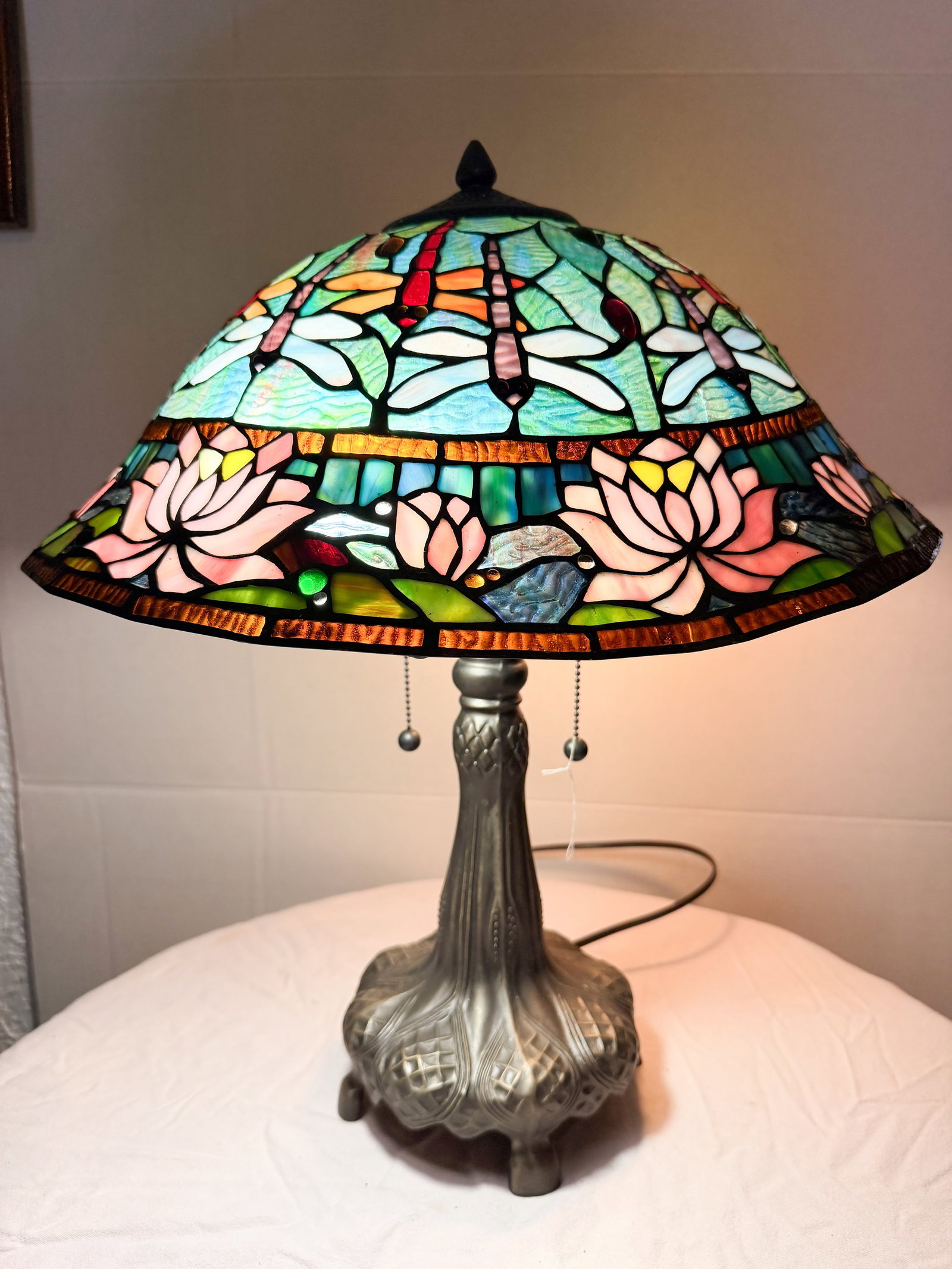 TIFFANY STYLE DRAGONFLY TABLE LAMP: table lamp featuring a repeating dragonfly and water lily motif set on a fluted cast metal base with a bronze-toned patina. The shade is constructed using the copper foil technique and incorporates