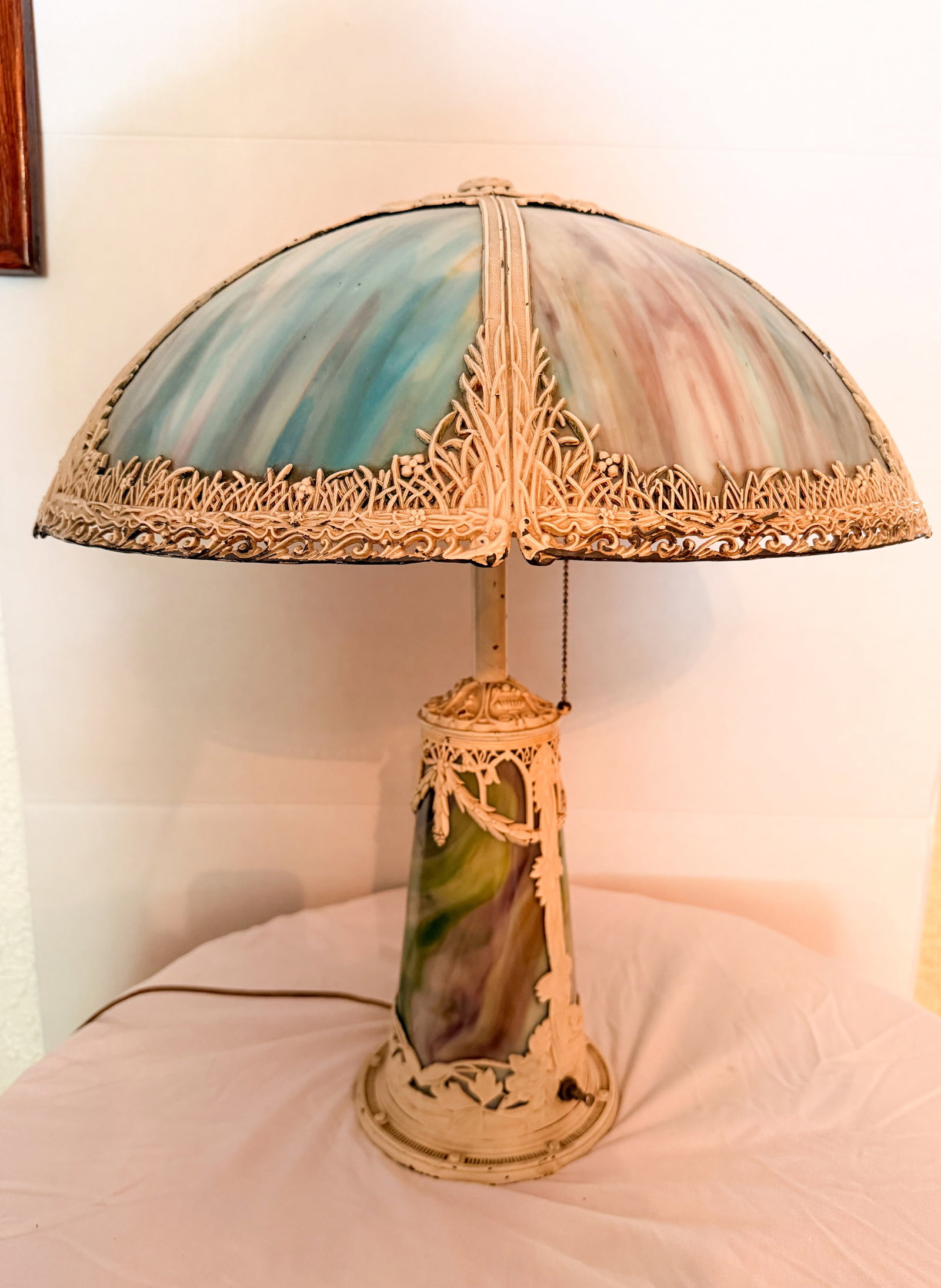 SLAG GLASS OVERLAY LAMP, CIRCA 1920: An early 20th-century table lamp featuring a six-panel curved slag glass shade with marbled aqua, mauve, and cream tones beneath an ornate wheat-stalk metal overlay, supported by a coordinating