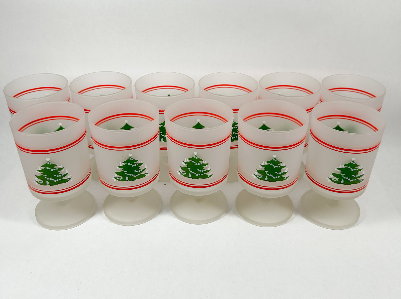 11 FROSTED CHRISTMAS TREE GOBLETS: A set of frosted glass goblets featuring screen-printed green Christmas tree motifs and red horizontal bands. The vessels are constructed with a cylindrical bowl, short stem, and circular foot.