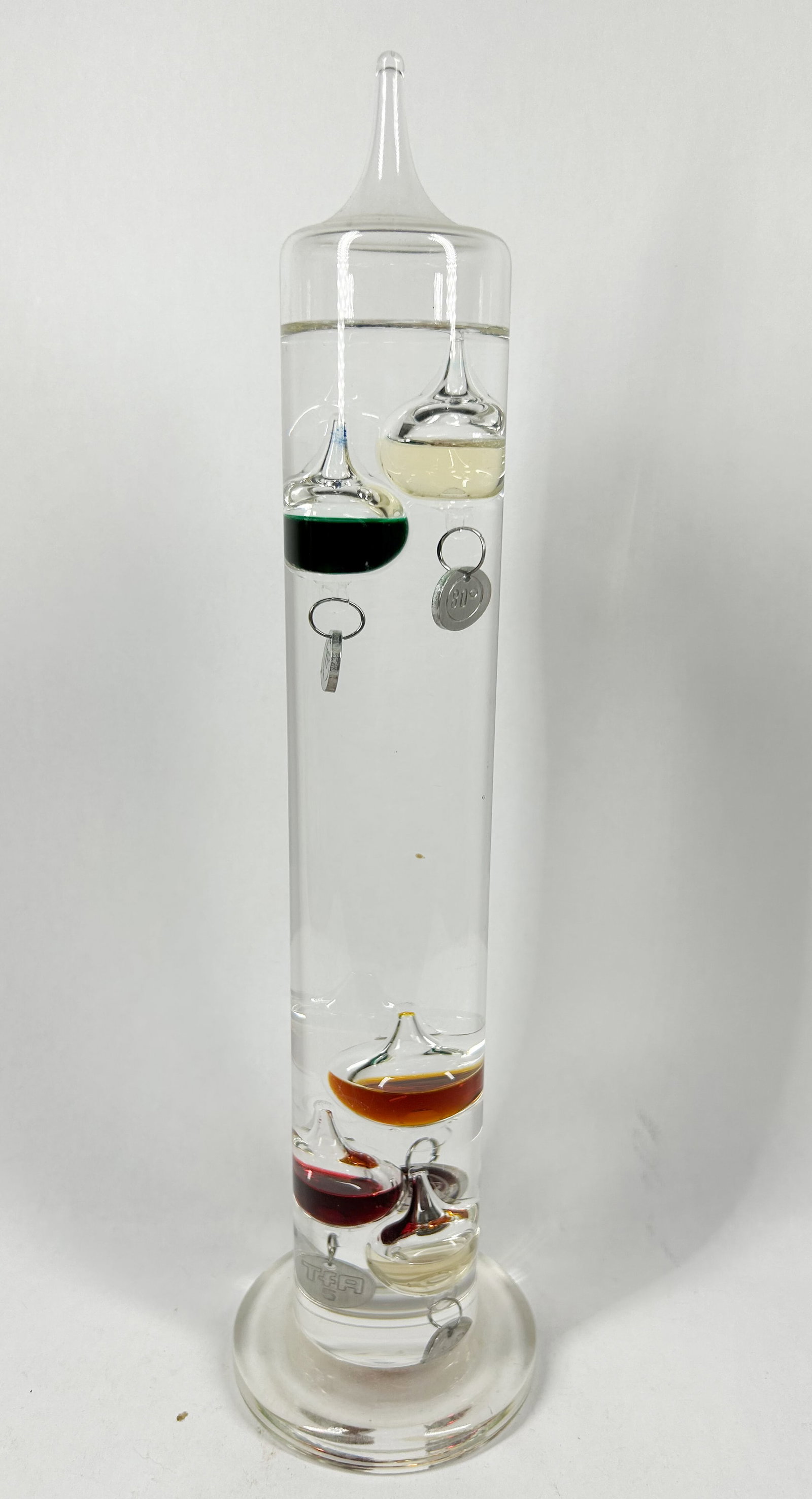 TFA DOSTMANN GLASS GALILEO THERMOMETER: Glass Galileo thermometer manufactured by TFA Dostmann featuring five internal spheres filled with multi-colored liquids. A stamped metal medallion on the lowest sphere bears the block letters TFA.
