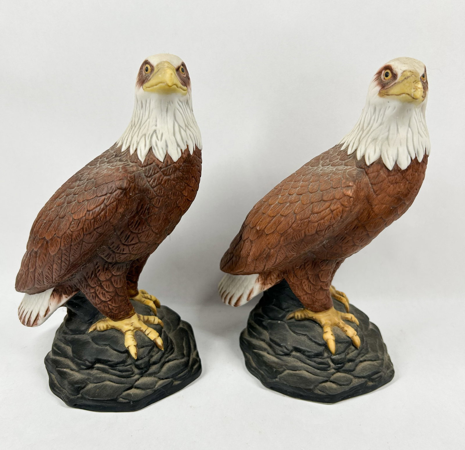 BRUEY PAIR PORCELAIN BALD EAGLES 1982: Bruey hand-painted bisque porcelain bald eagle figurines. Each figurine features an incised script signature on the reverse of the rocky base reading 'Bruey'.