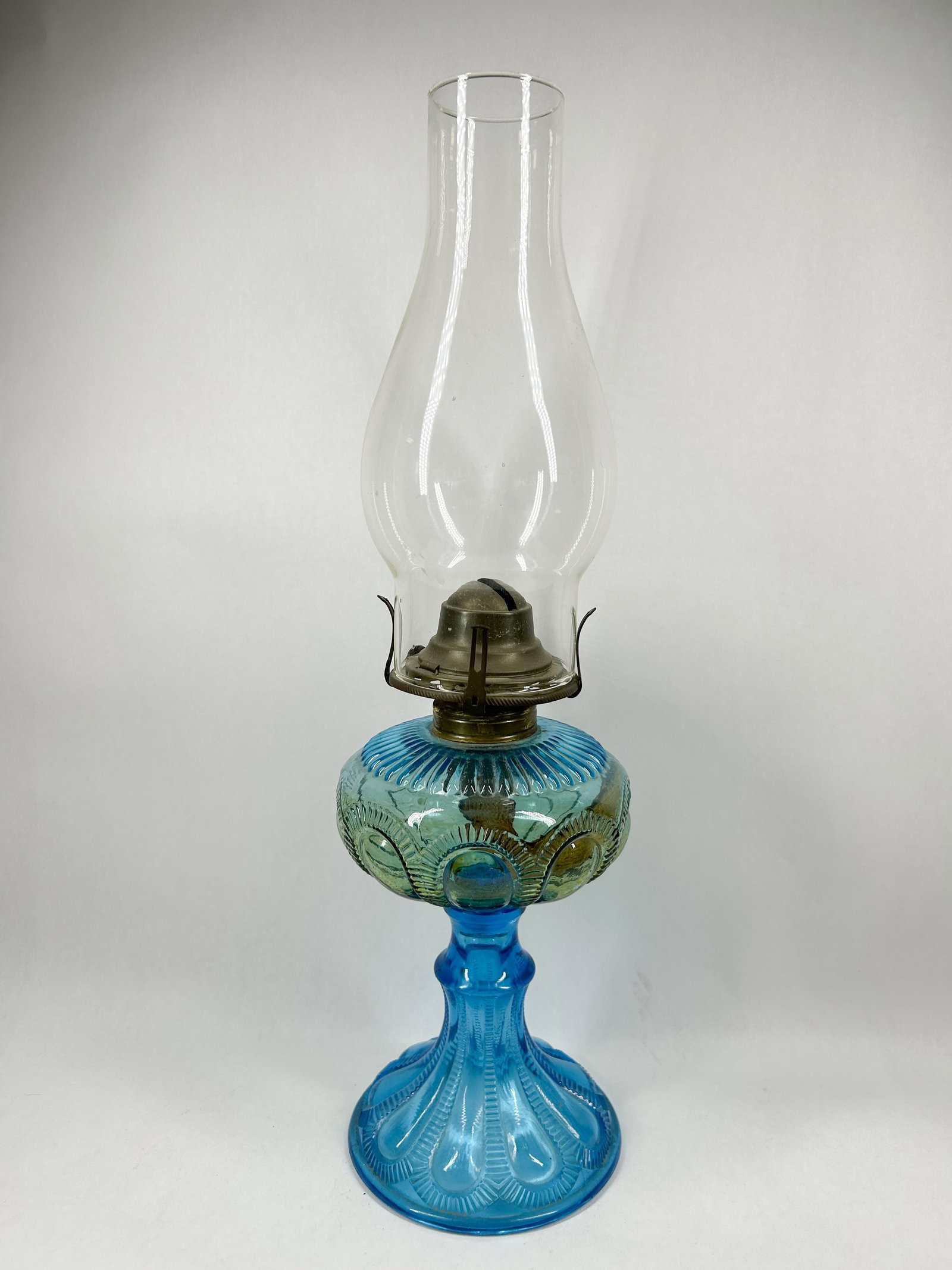 BLUE GLASS KEROSENE LAMP: Molded glass kerosene lamp featuring a teal blue font with a circular bullseye and fan motif joined to a cobalt blue fluted pedestal base. The lamp is equipped with a metal burner assembly featuring