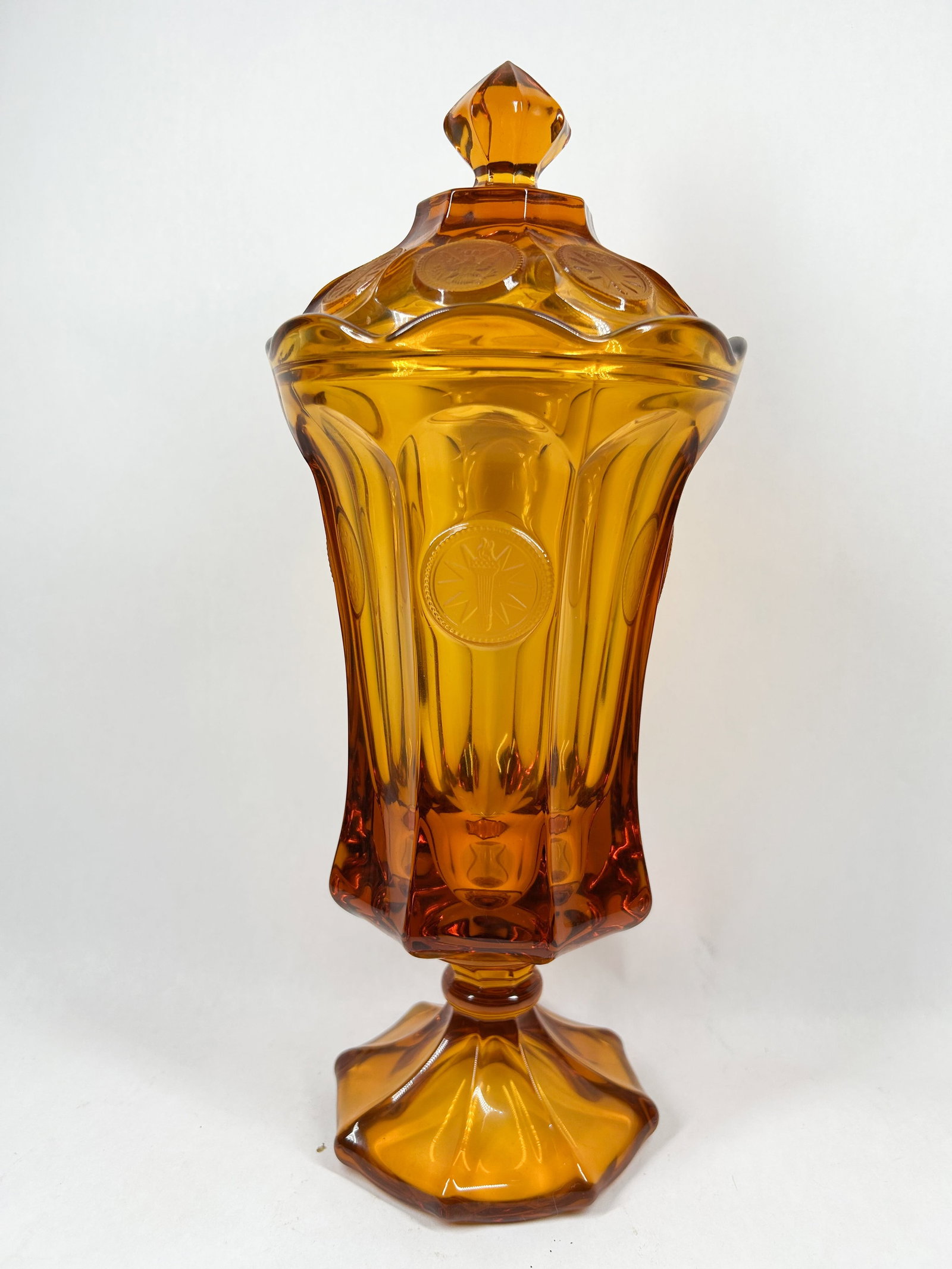 FOSTORIA AMBER COIN GLASS JAR, 1960S: Fostoria Glass Company pressed amber glass apothecary jar in the Coin Glass pattern. The vessel features fluted panels and commemorative coin medallions depicting a heraldic eagle and a sunray torch.