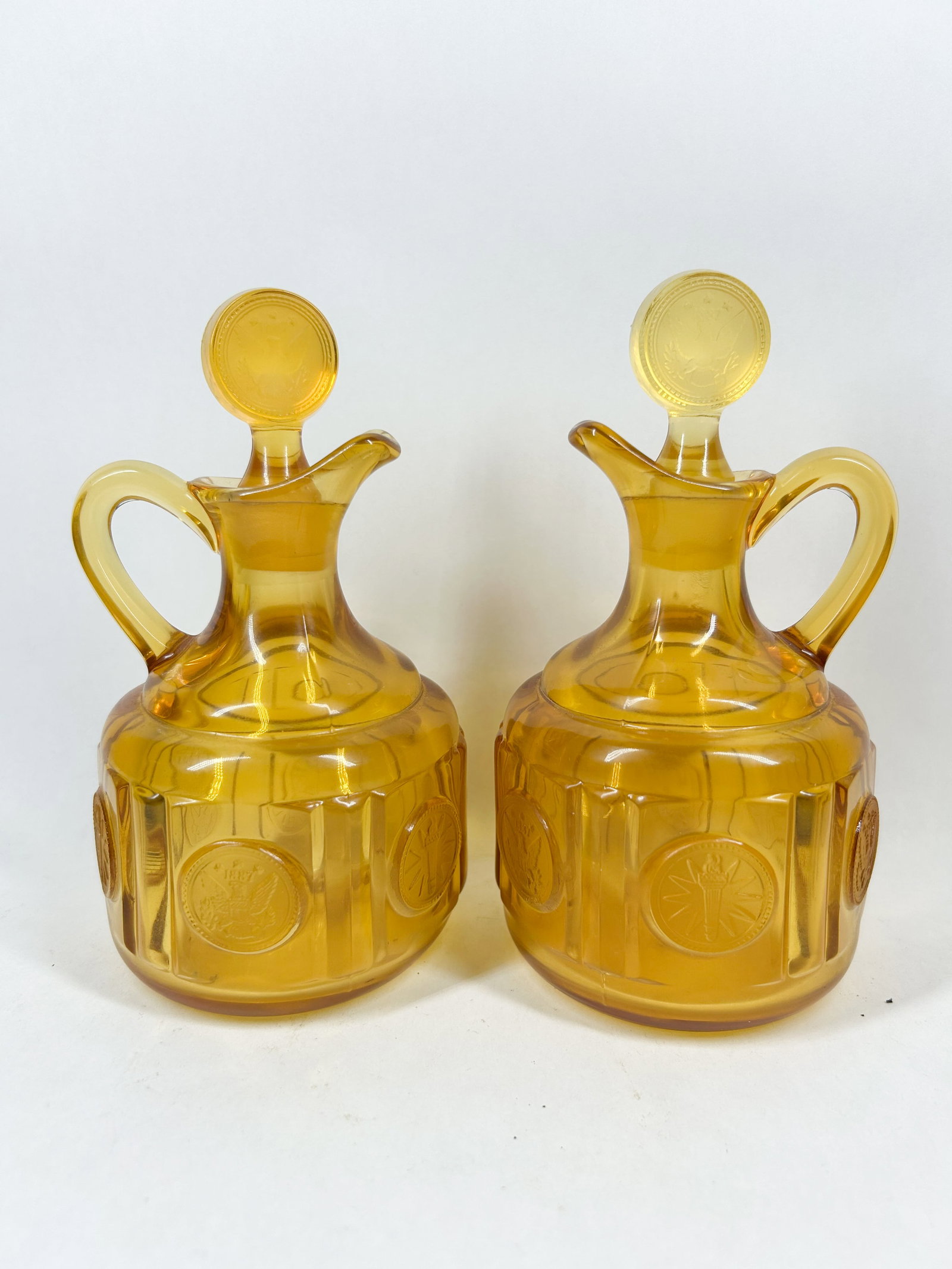 AMBER COIN GLASS CRUETS, C. 1960: A pair of amber-colored pressed glass cruets featuring the Coin Glass pattern. Each vessel is decorated with molded medallions dated 1887 and accompanied by matching eagle-motif disc stoppers.