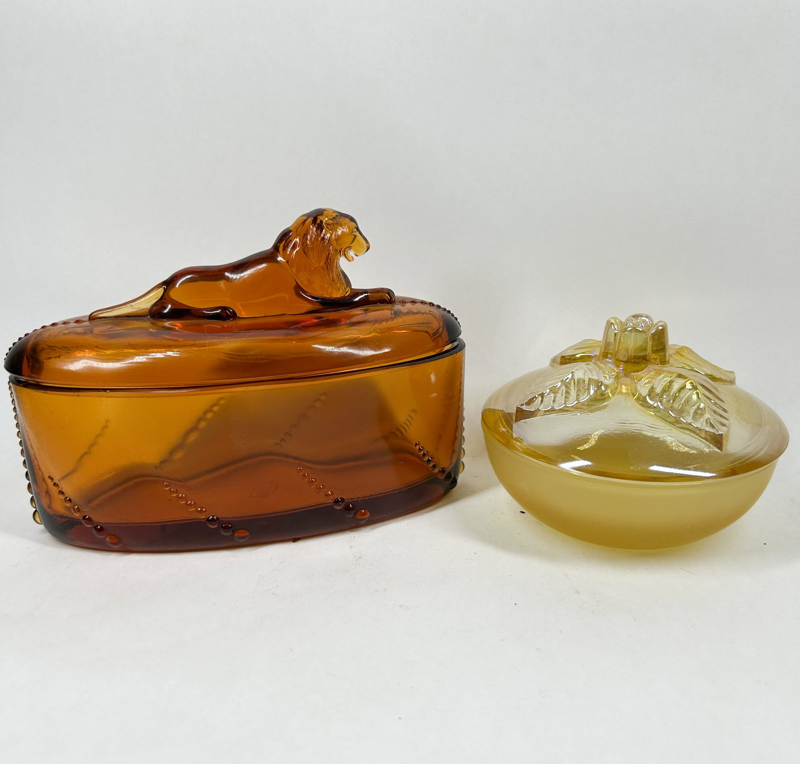 TWO VINTAGE PRESSED GLASS TRINKET BOXES, 20TH CENTURY: Two vintage pressed glass trinket boxes. The deep amber oval box features a reclining lion finial and beaded swag motifs, while the iridescent yellow round box features a rose finial and a satin finis