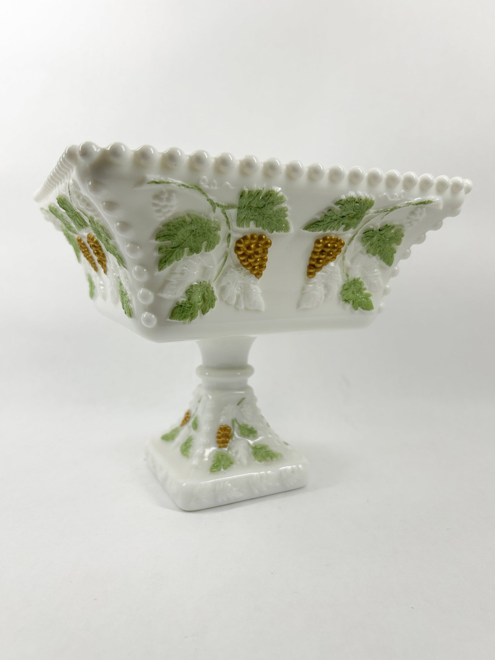 PAINTED MILK GLASS SQUARE COMPOTE: A vintage square milk glass pedestal compote featuring a beaded rim and relief-molded grapevine decoration. The exterior grapes are accented in gold-tone metallic paint with green painted leaves and