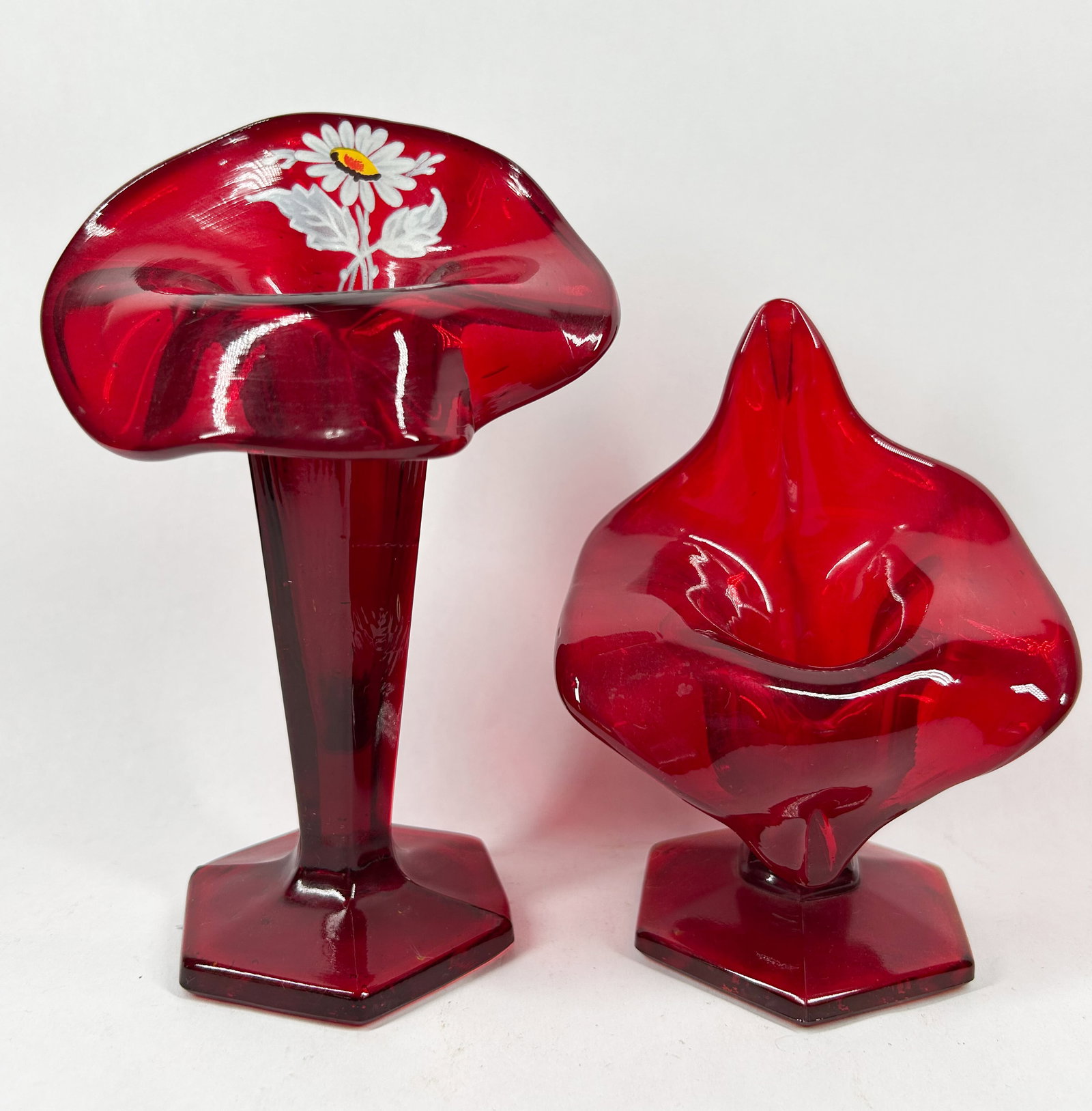 VINTAGE RUBY GLASS VASE PAIR: A pair of hand-blown ruby glass vases featuring pressed hexagonal pedestal bases. The taller vessel displays a Jack-in-the-pulpit form with a hand-painted floral motif, while the smaller piece