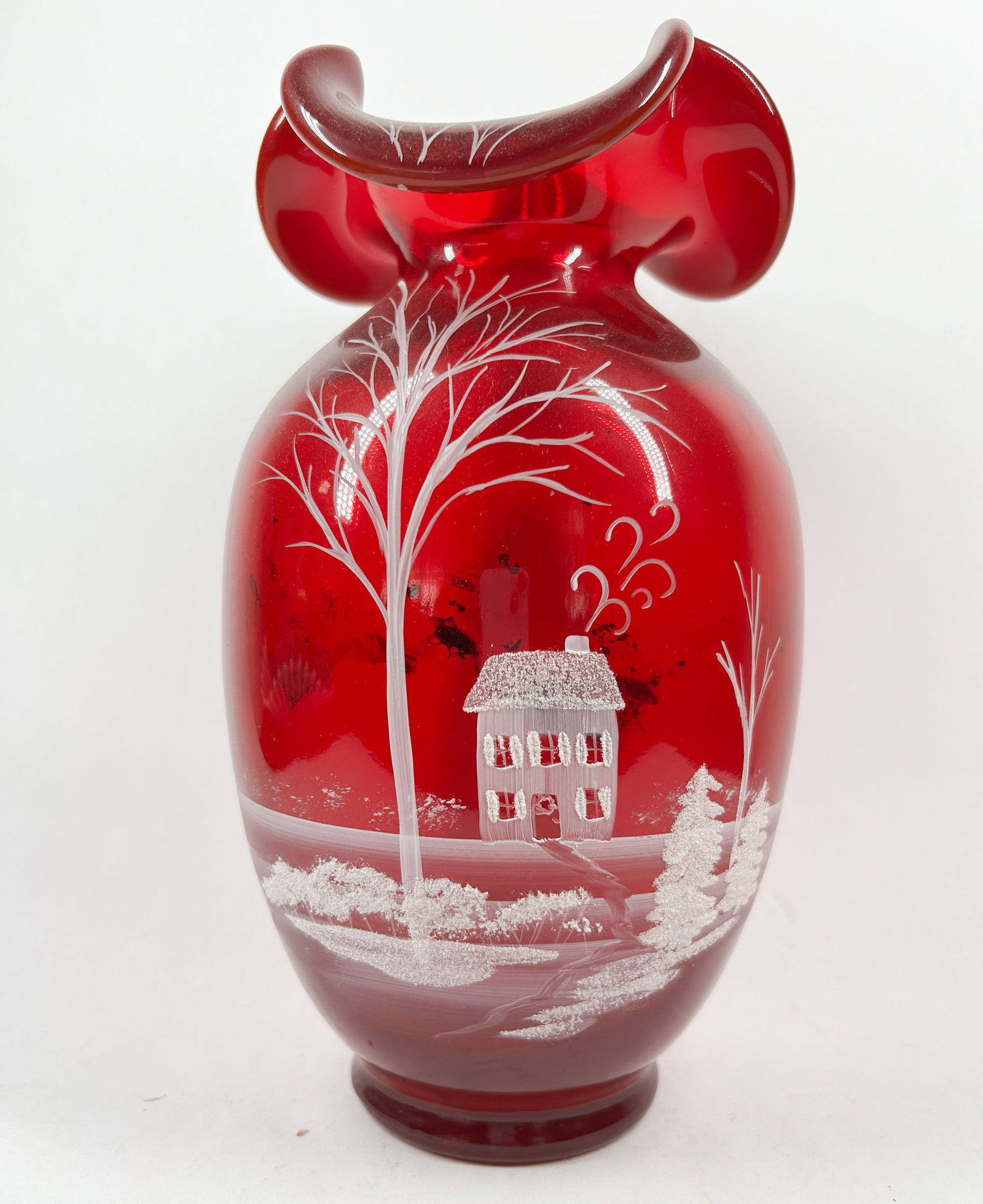 FENTON RUBY RED GLASS WINTER VASE: Hand-blown ruby red glass vase featuring a ruffled tri-corner rim and a hand-painted white enamel winter landscape. The scene depicts a cottage and trees with textured enamel snow above a polished
