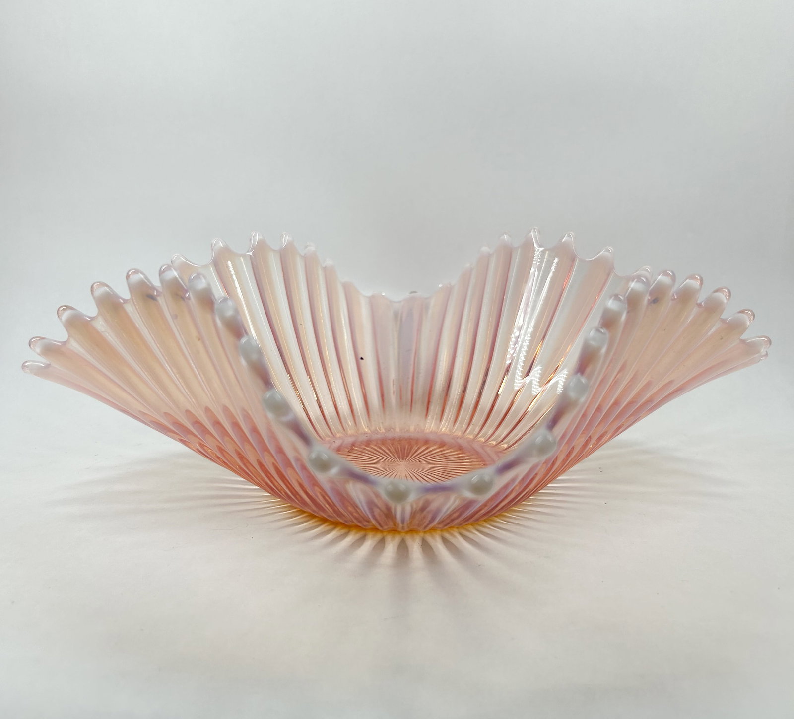 FENTON PINK OPALESCENT GLASS RUFFLED BOWL: Mid-20th century opalescent glass bowl featuring a translucent pink body and a white opalescent, ruffled rim. The vessel is decorated with radiating vertical ribs and a central sunburst pattern on