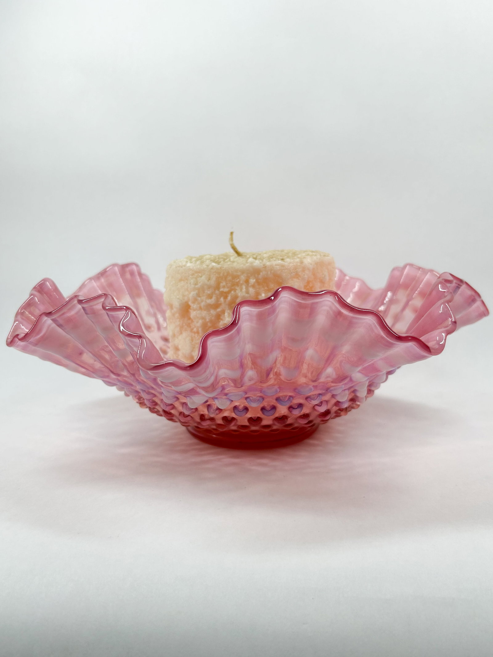 FENTON CRANBERRY OPALESCENT HOBNAIL GLASS BOWL: A vintage glass bowl featuring a cranberry opalescent finish and a hobnail pattern. The piece is finished with a deeply ruffled, crimped rim and is accompanied by a textured pillar candle. Candle is