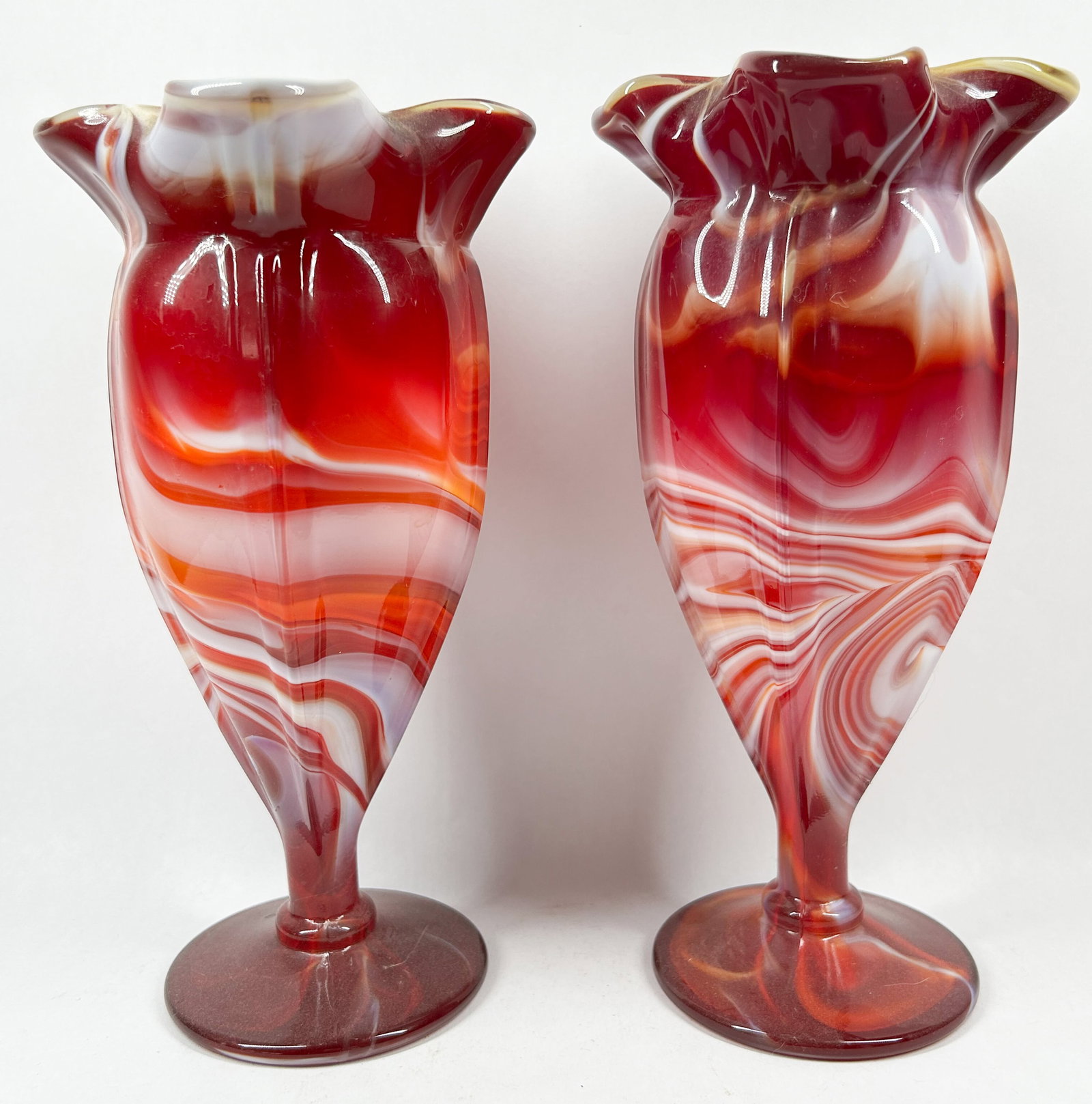 PAIR OF RED SLAG GLASS VASES, IMPERIAL: A pair of opaque slag glass vases characterized by a red and white marbled pattern. Each vessel features a six-point ruffled rim above a bulbous body and a solid pedestal foot.
